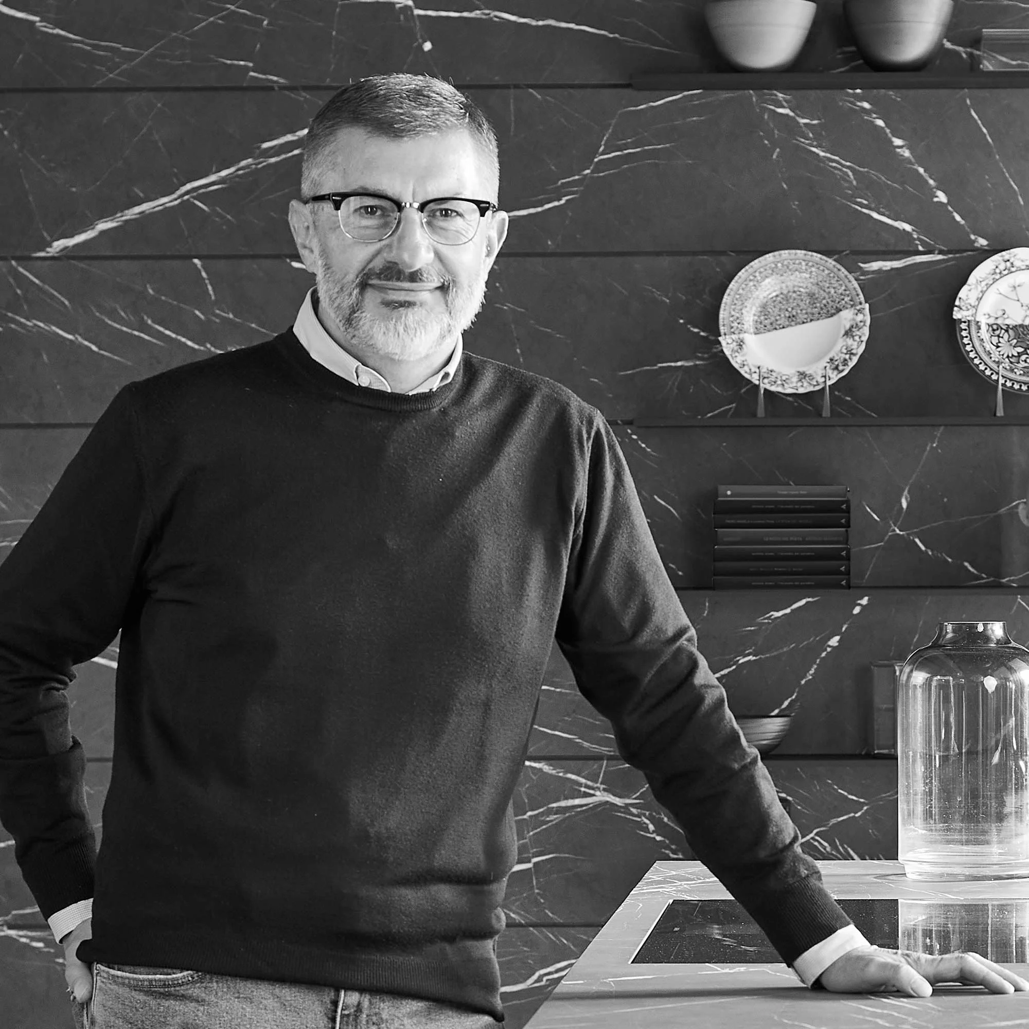 Uomo con capelli corti e barba, indossa occhiali, maglione nero e camicia bianca, si appoggia a un tavolo in una cucina moderna.