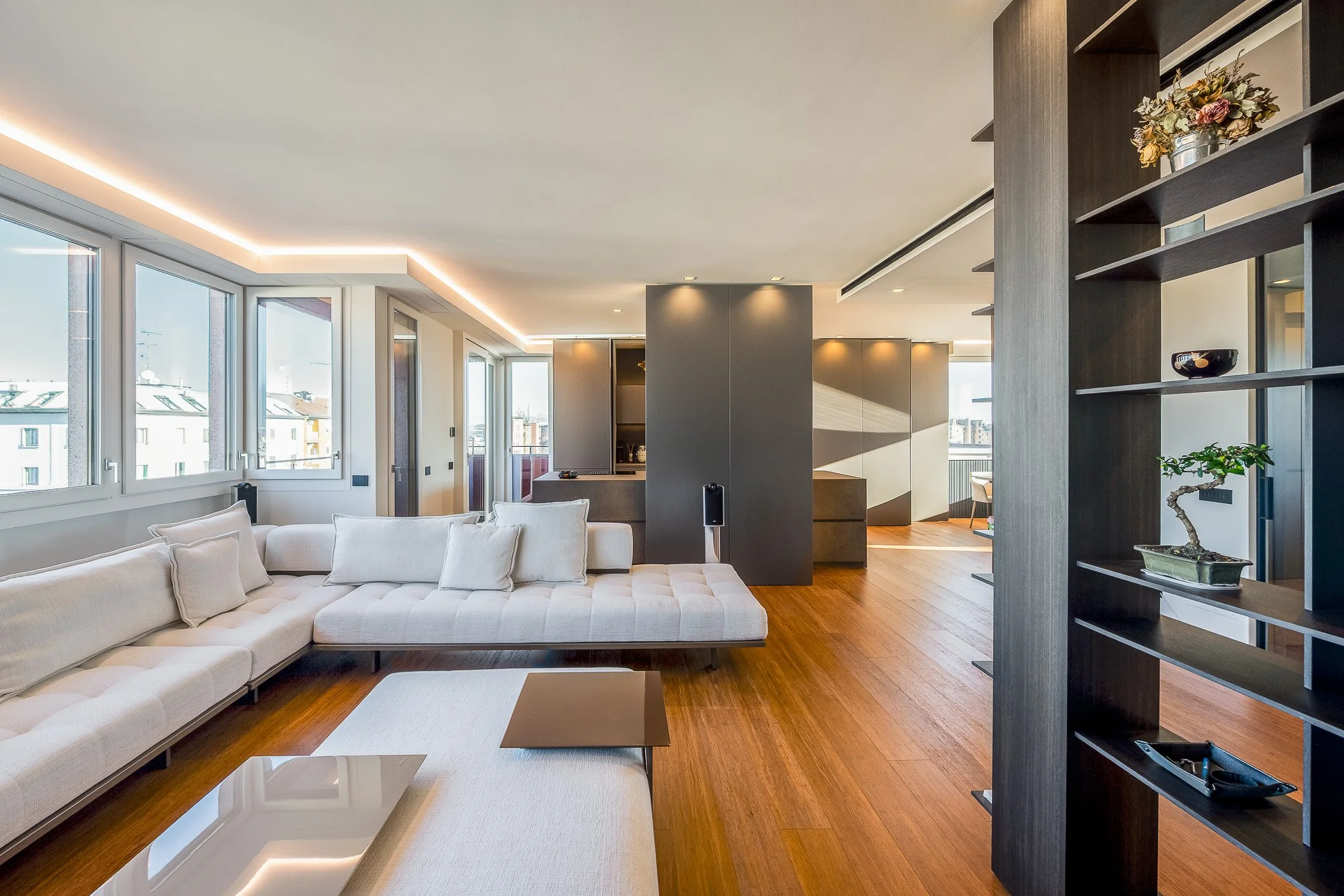 Interno di un soggiorno moderno con grandi finestre, divano bianco, mobili in legno scuro, scaffali decorativi e dettagli di design.