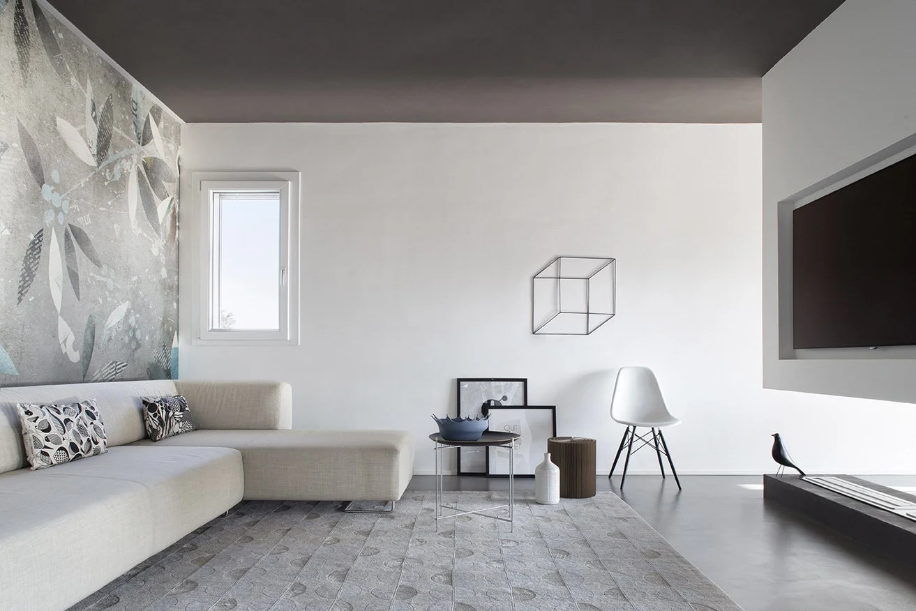 Interno di soggiorno moderno con parete decorata, divano chiaro, vari quadri, sedia bianca, tavolino e televisore.