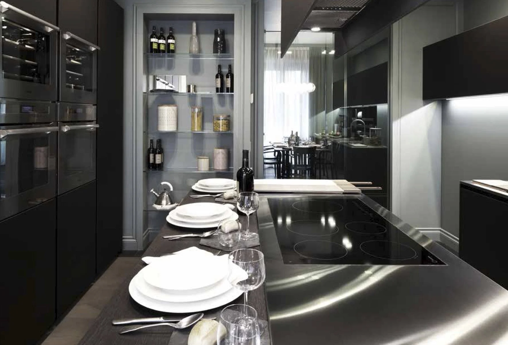 Cucina moderna con piano cottura in acciaio inox, tavolo con piatti, bicchieri e bottiglia di vino, scaffale con bottiglie e contenitori, area pranzo visibile attraverso una porta a vetri.