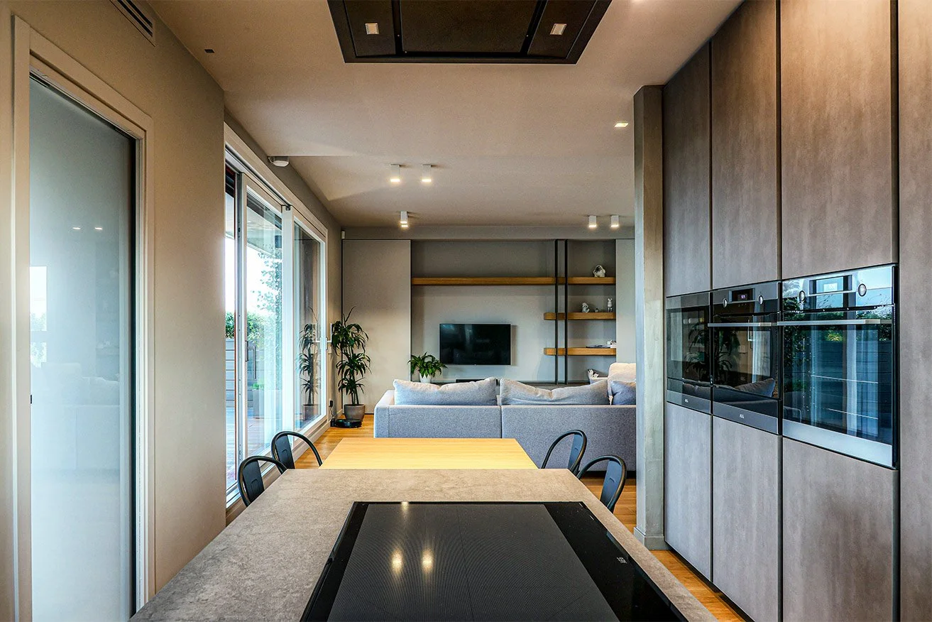 Interno di una moderna zona living e cucina con tavolo ovale, sedie nere, pareti con grandi finestre, divano grigio, televisore, mensole di legno e cucina con armadietti grigi e forno.