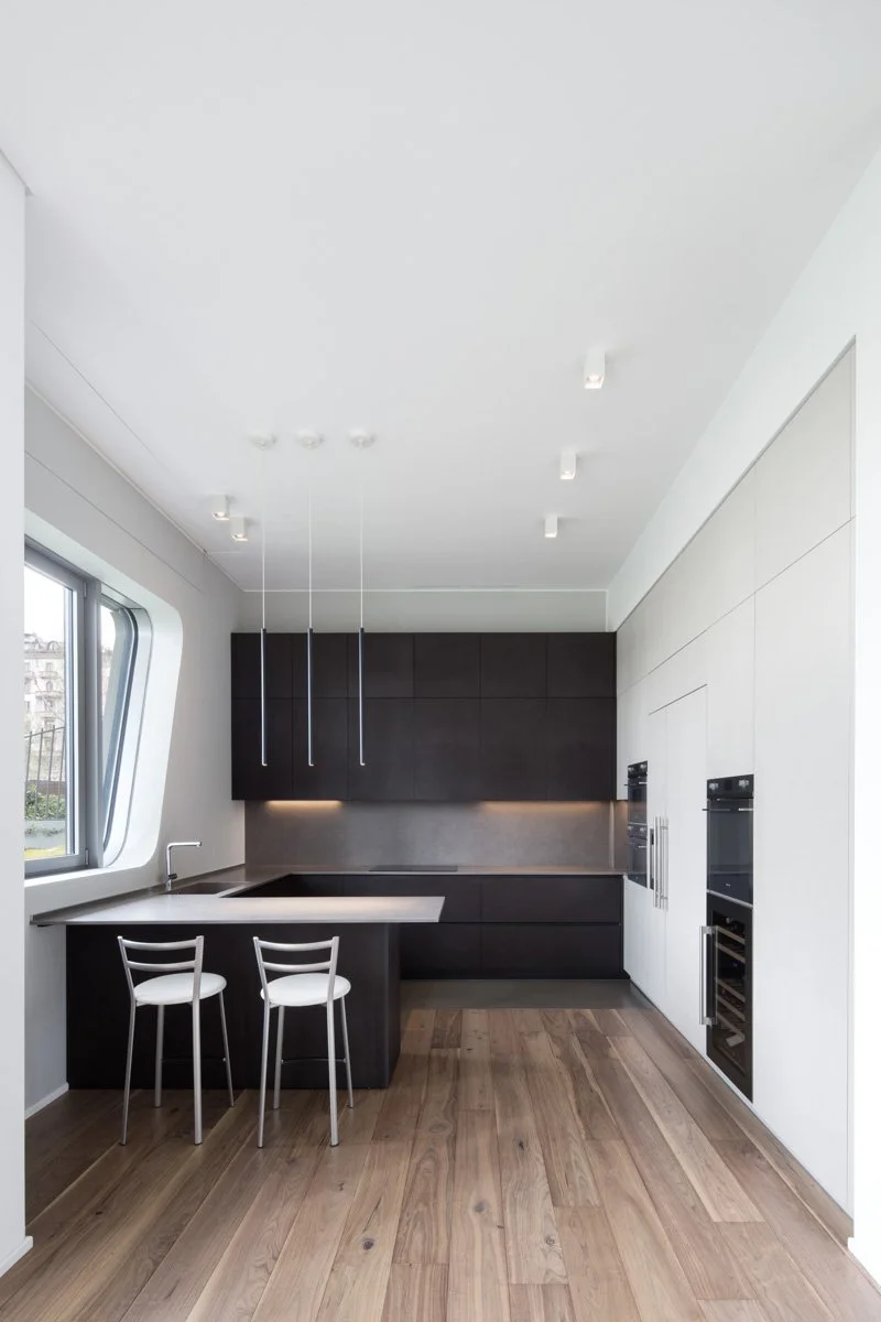 Cucina moderna con mobili neri e bianco, due sedie bianche e un piano di lavoro in acciaio, pareti lisce e pavimento in legno, finestra laterale.