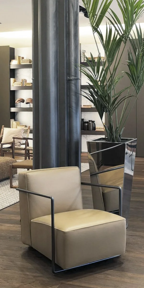 Sedia moderna in pelle beige con braccioli in metallo, grande pianta verde in vaso decorativo e libreria con libri e oggetti in arredamento d'interni.