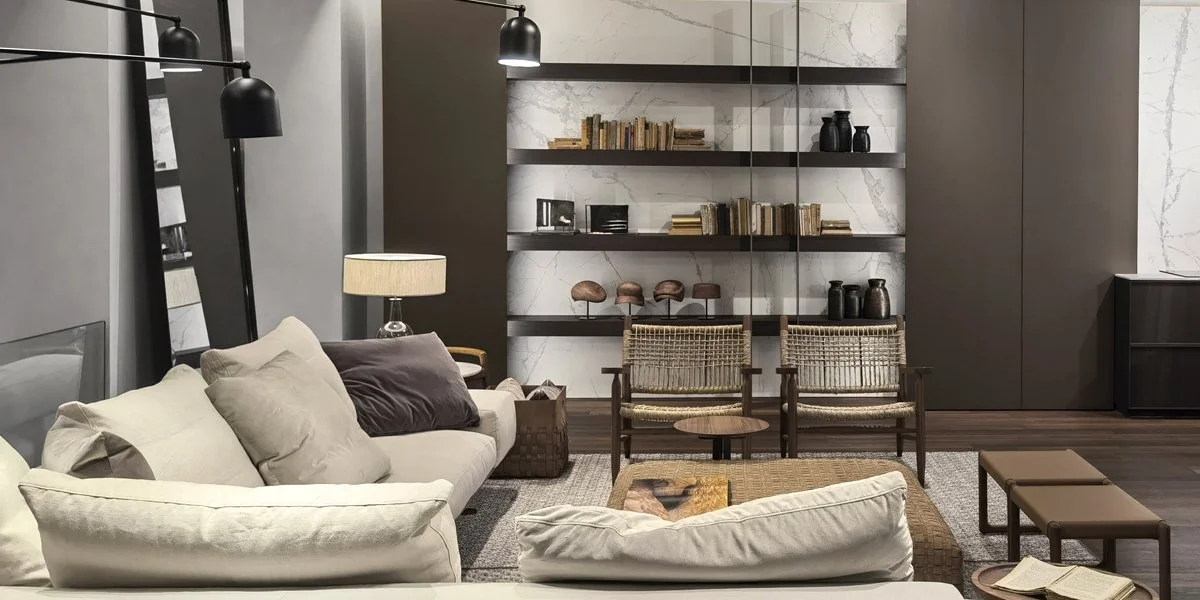 Soggiorno elegante con divano beige, poltrone di legno, libreria con libri e oggetti decorativi, lampade nere e una lampada da tavolo. Pareti in marmo e mobili moderni.