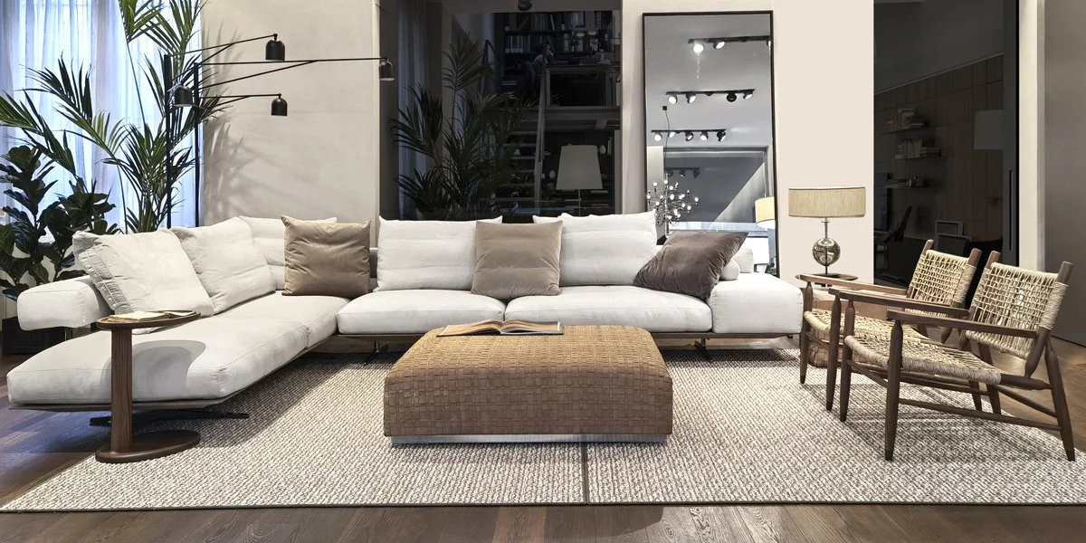 Soggiorno moderno con grande divano bianco, pouf marrone, quattro sedie in legno con seduta in paglia, lampada da tavolo, lampade da parete e grandi piante verdi.