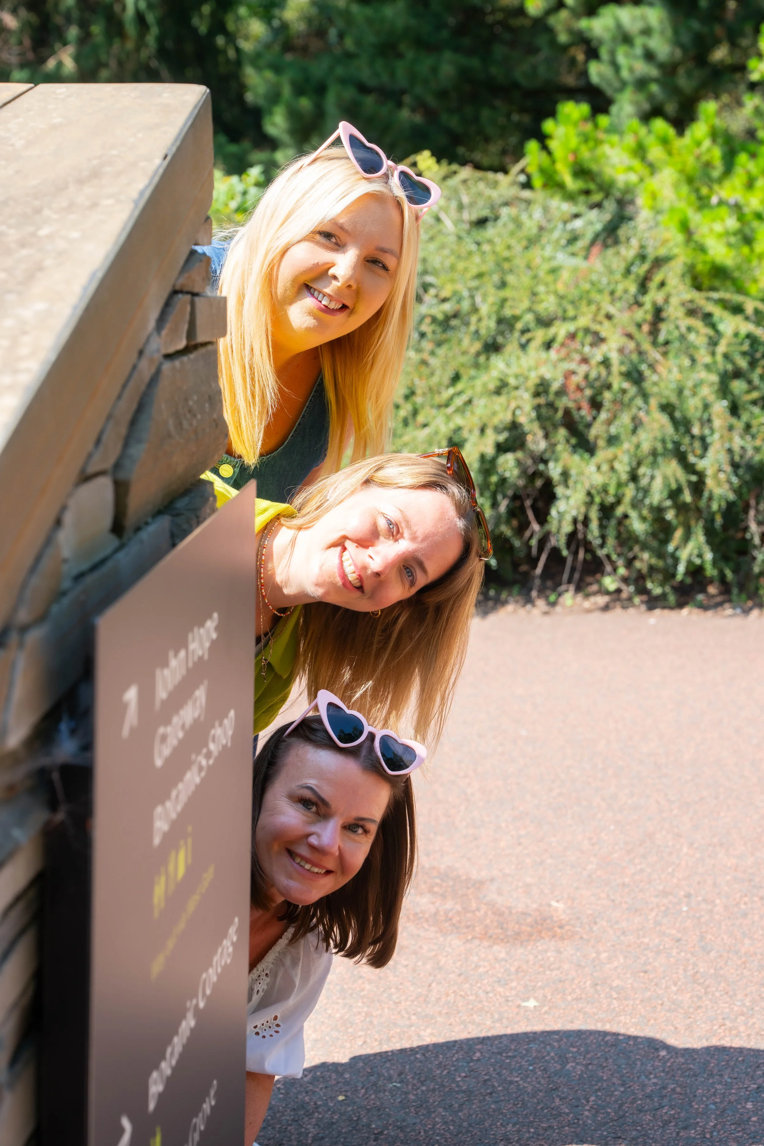 Four smiling women peeking out from behind a wooden sign outdoors on a sunny day, with trees in the background. They are wearing sunglasses and casual clothing.