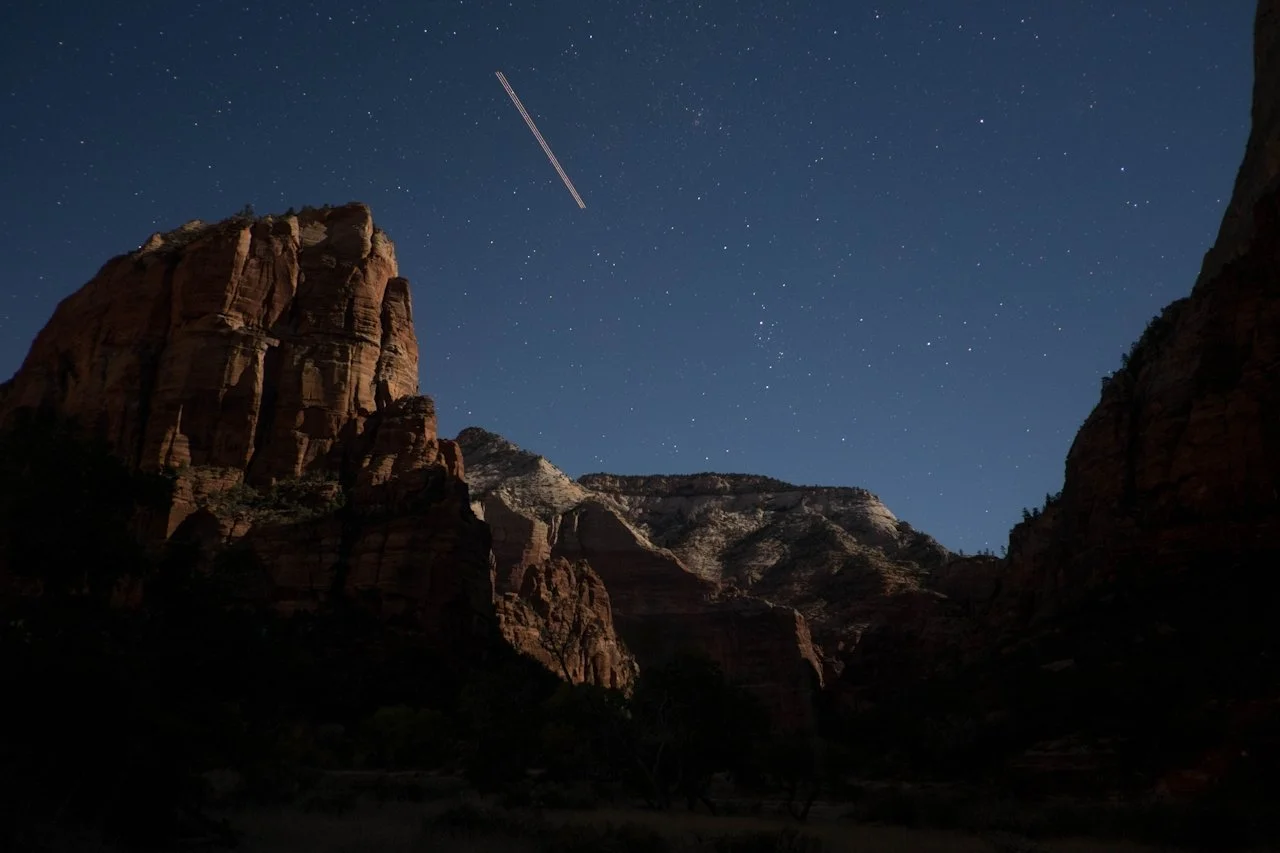 Night sky over a canyon with stars and a streak of light from a passing airplane.