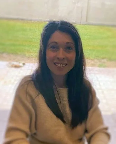 Smiling woman with long dark hair, wearing a beige sweater, sitting outdoors near a grassy area and a concrete wall.