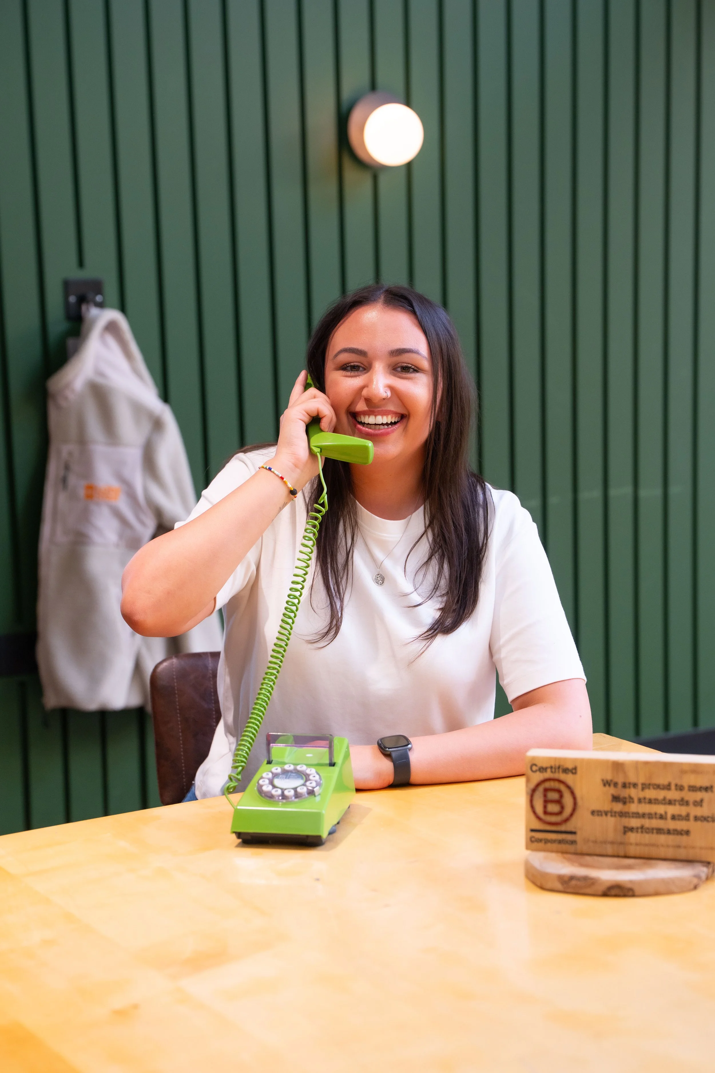 A young woman with dark hair and a nose piercing smiling while talking on a vintage green rotary phone. She is wearing a white t-shirt, a necklace, a colorful bracelet, and a watch. The background features green vertical wall panels, a light fixture, a coat hanging on a hook, and a wooden block with text. She is sitting at a wooden table with a green desk phone and a small display case.