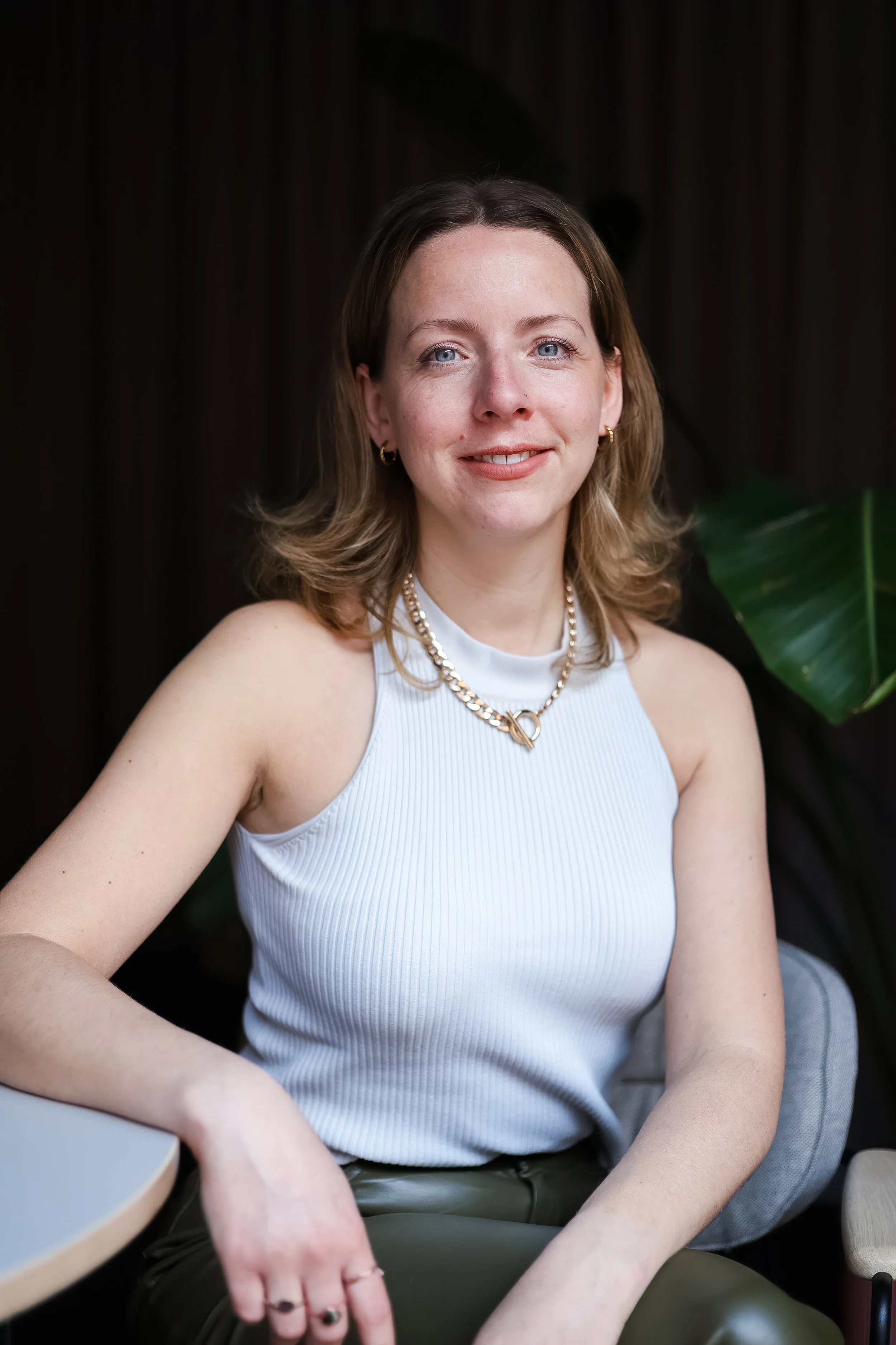 A woman with shoulder-length brown hair, blue eyes, wearing earrings, a pearl necklace, and a white sleeveless top, smiling softly, sitting with her left arm resting on a surface, and a large green plant is visible in the background.