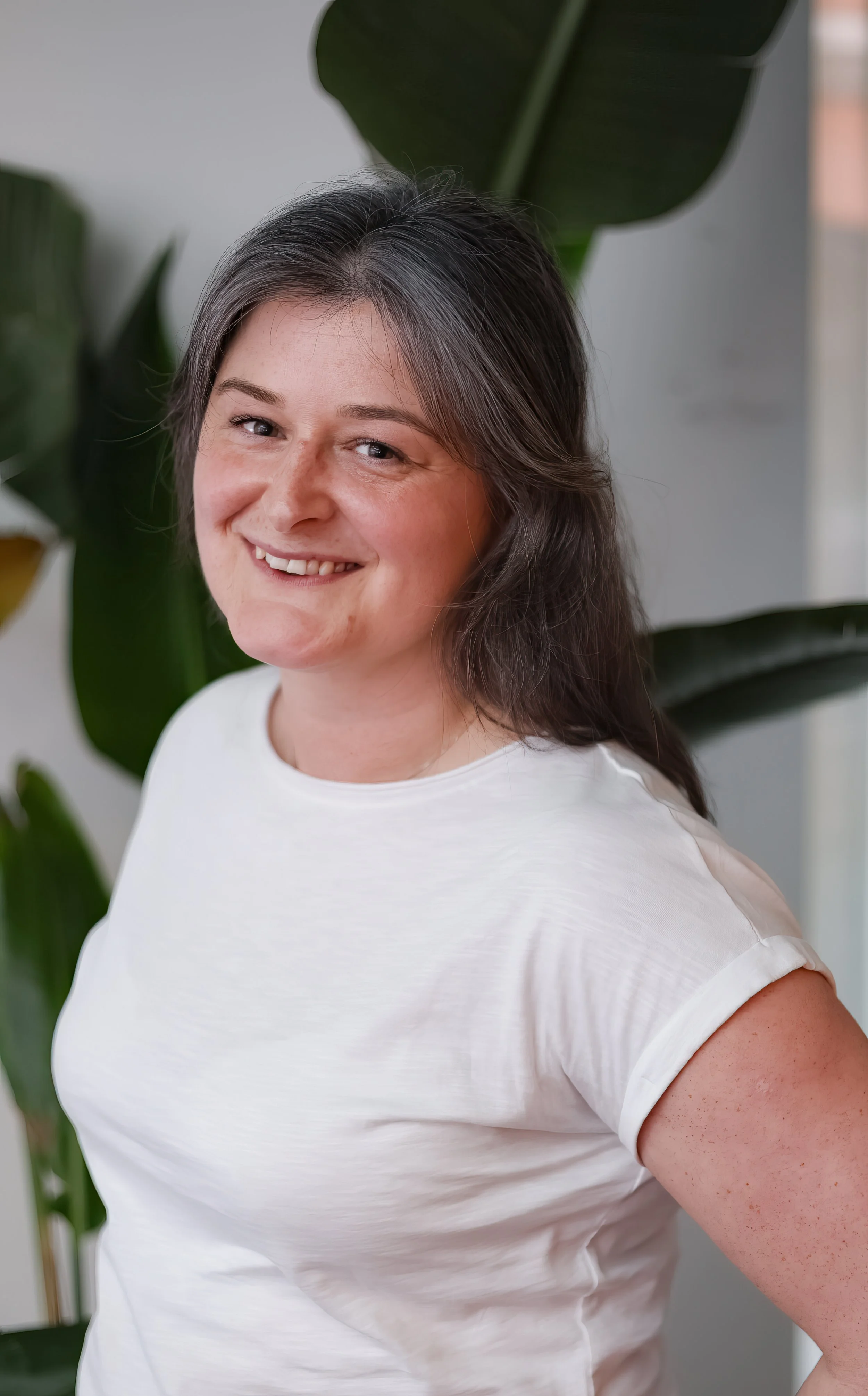 Close-up of a smiling woman with shoulder-length gray hair, wearing a white T-shirt, standing in front of large green leaves.