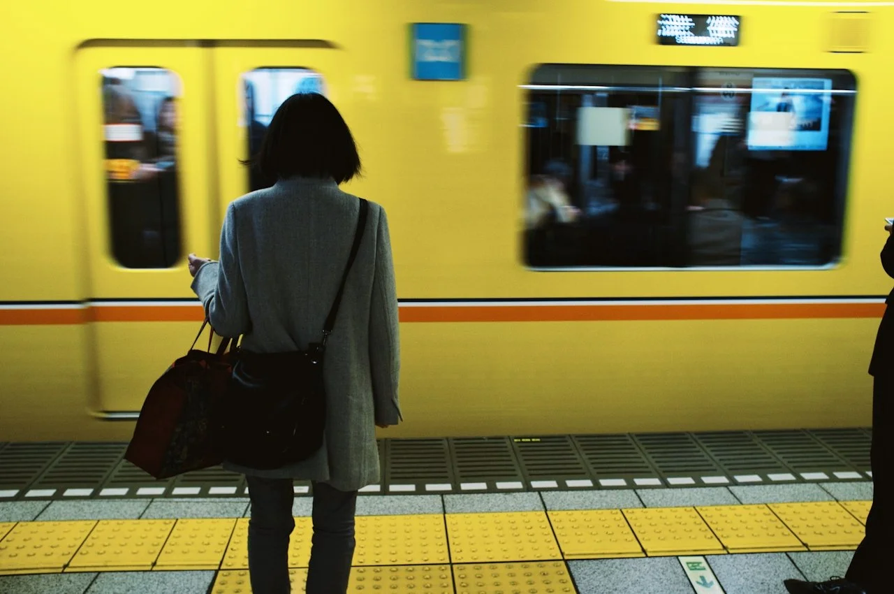 A woman with a shoulder bag standing on a yellow safety line at a subway station, waiting for the train to pass. A yellow train is arriving behind her.
