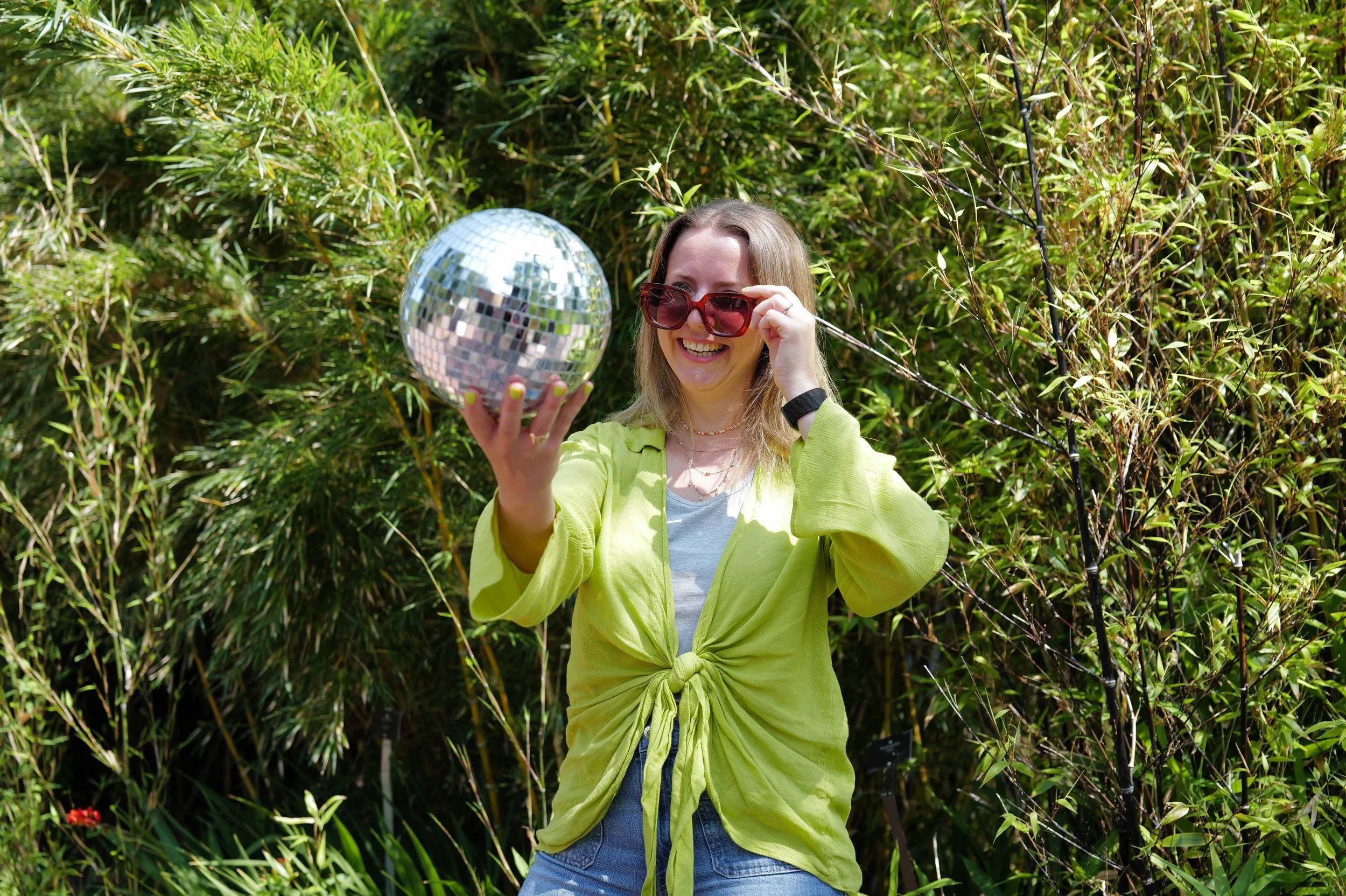 A woman smiling and holding a disco ball outdoors in front of green bushes, wearing red sunglasses and a green jacket.