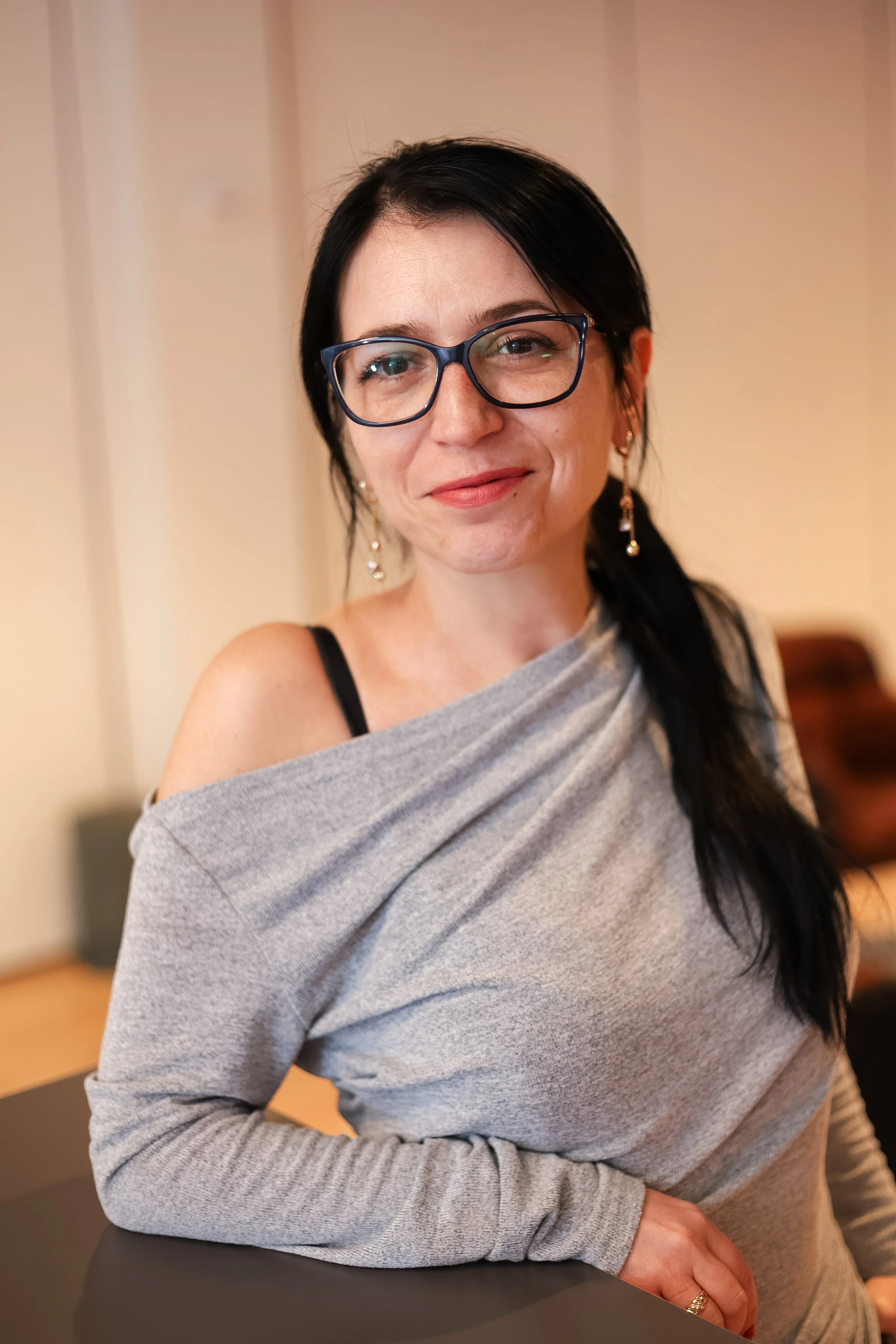 A woman with black hair, glasses, and earrings is sitting at a table, wearing a gray off-shoulder top. She has a slight smile and is looking slightly to the side.