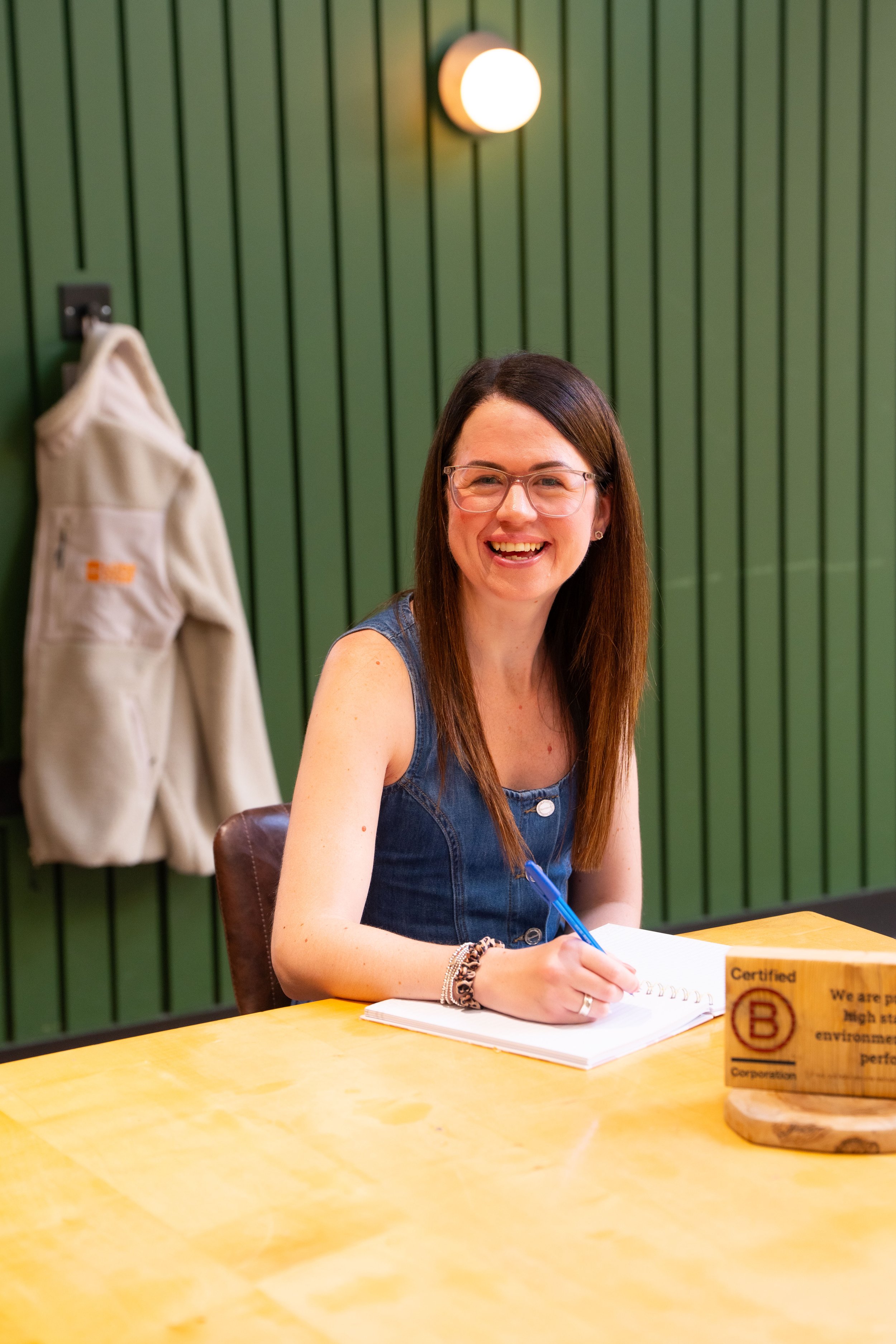 A smiling woman with glasses and long brown hair, wearing a denim sleeveless top, sitting at a yellow table with a notebook and blue pen, in front of a green paneled wall with a wall light and a beige jacket hanging on a hook.