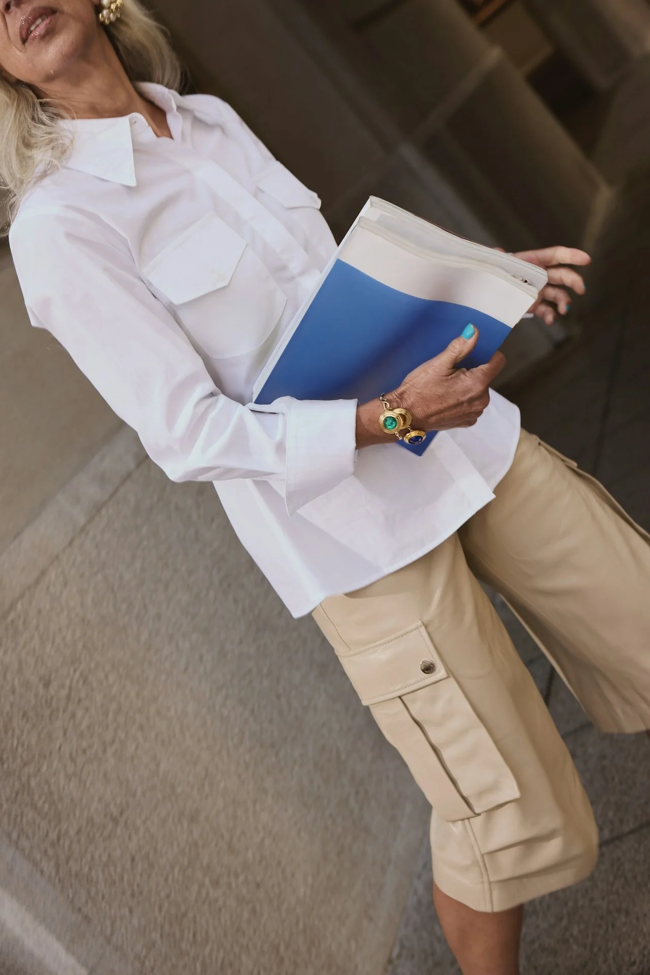 Woman standing indoors, holding a blue and white book or folder, wearing a white shirt and beige cargo shorts, with blonde hair and colorful jewelry.