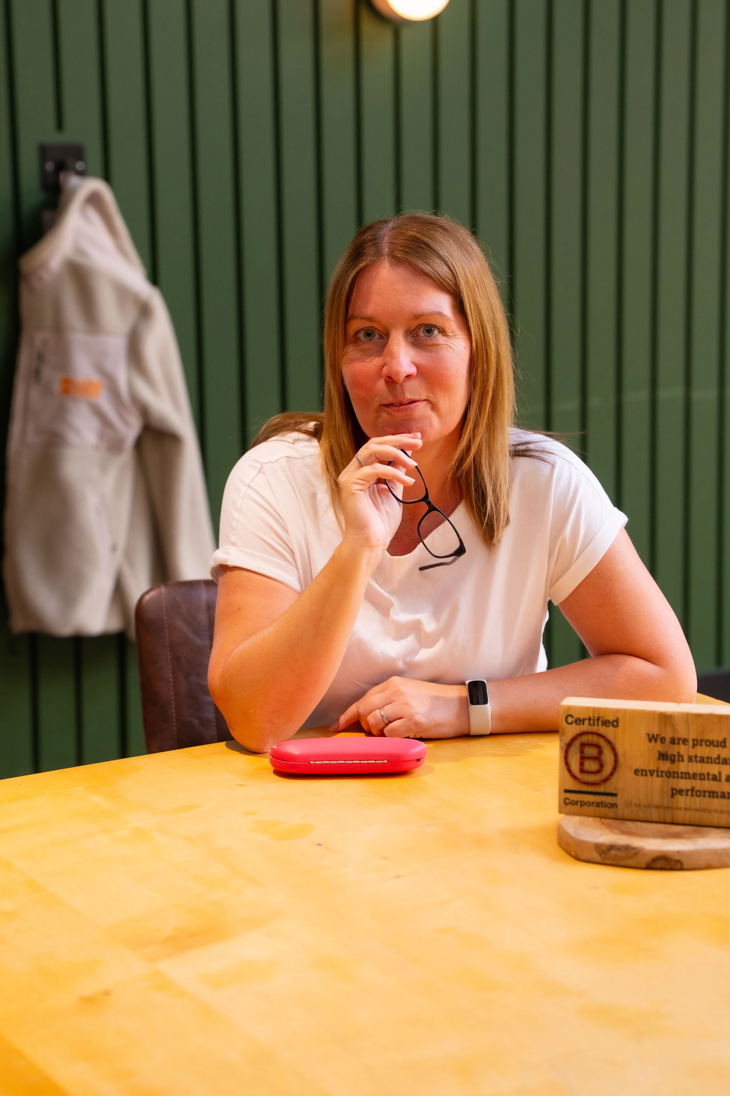 A woman with reddish-brown hair wearing a white t-shirt, holding black-rimmed glasses, sitting at a yellow table with a pink case and a wooden block with certification information. The background features a green vertical panel wall and a hanging light.