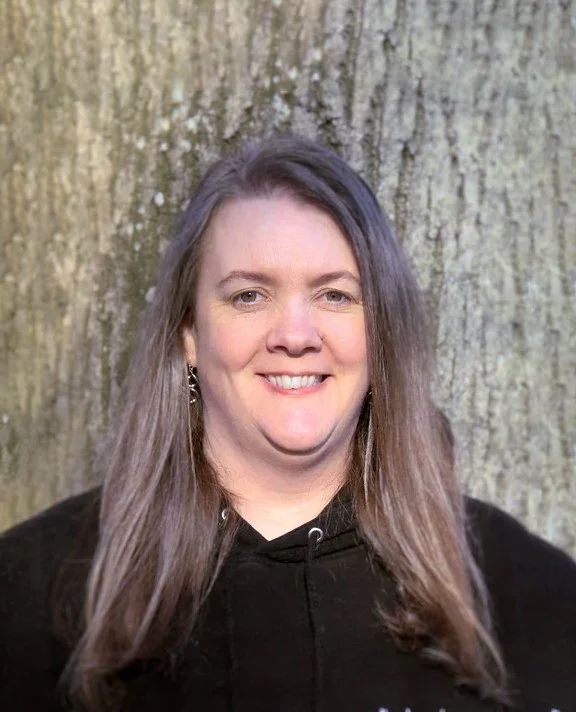 A woman with long brown hair and earrings smiling in front of a large tree trunk.