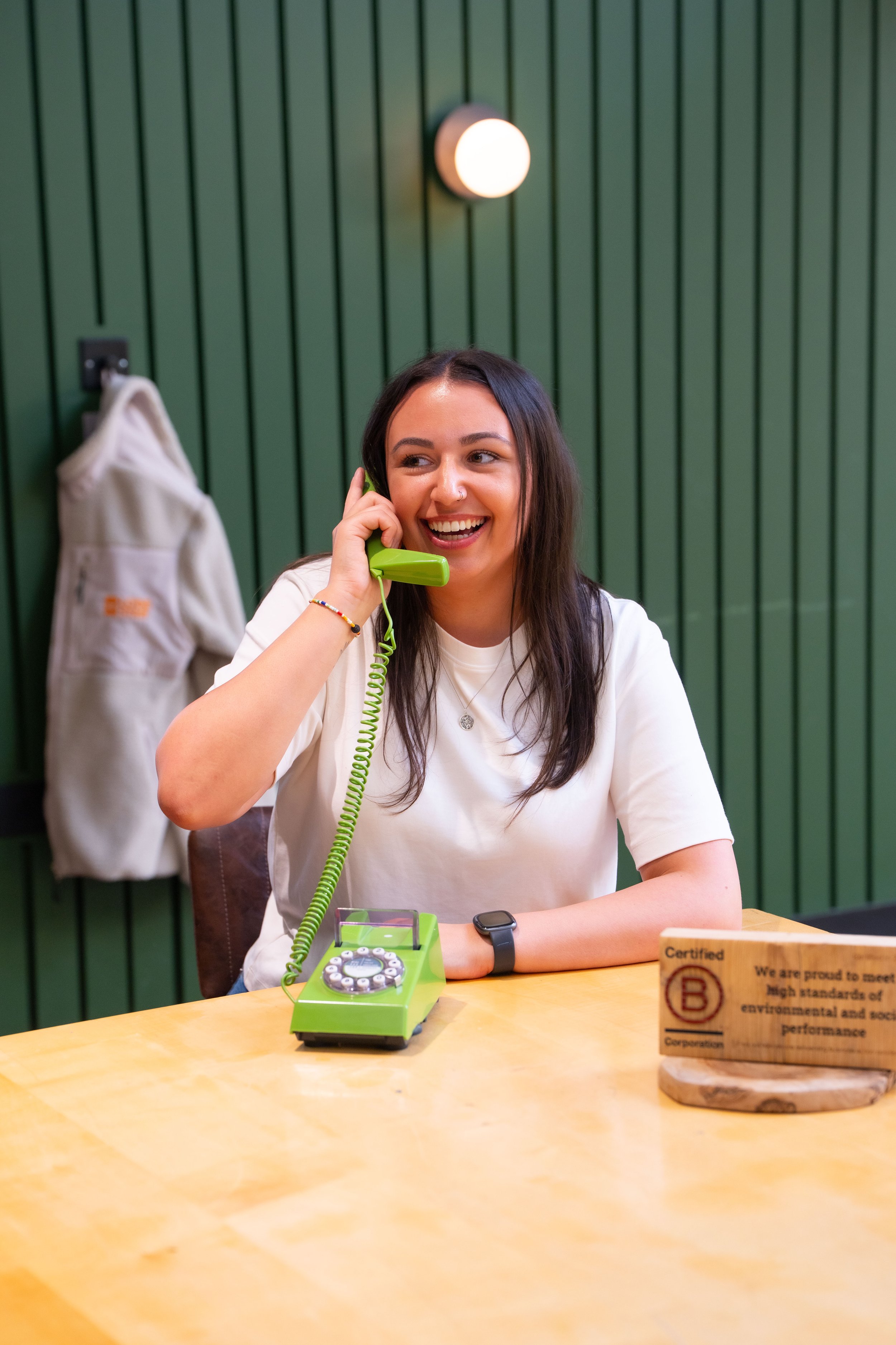 A young woman with long dark hair smiling and talking on a green vintage rotary telephone at a wooden table. She is wearing a white t-shirt and a black fitness tracker on her left wrist. In the background, there is a green wall with vertical wooden panels, a wall-mounted light, and a beige backpack hanging on a hook.