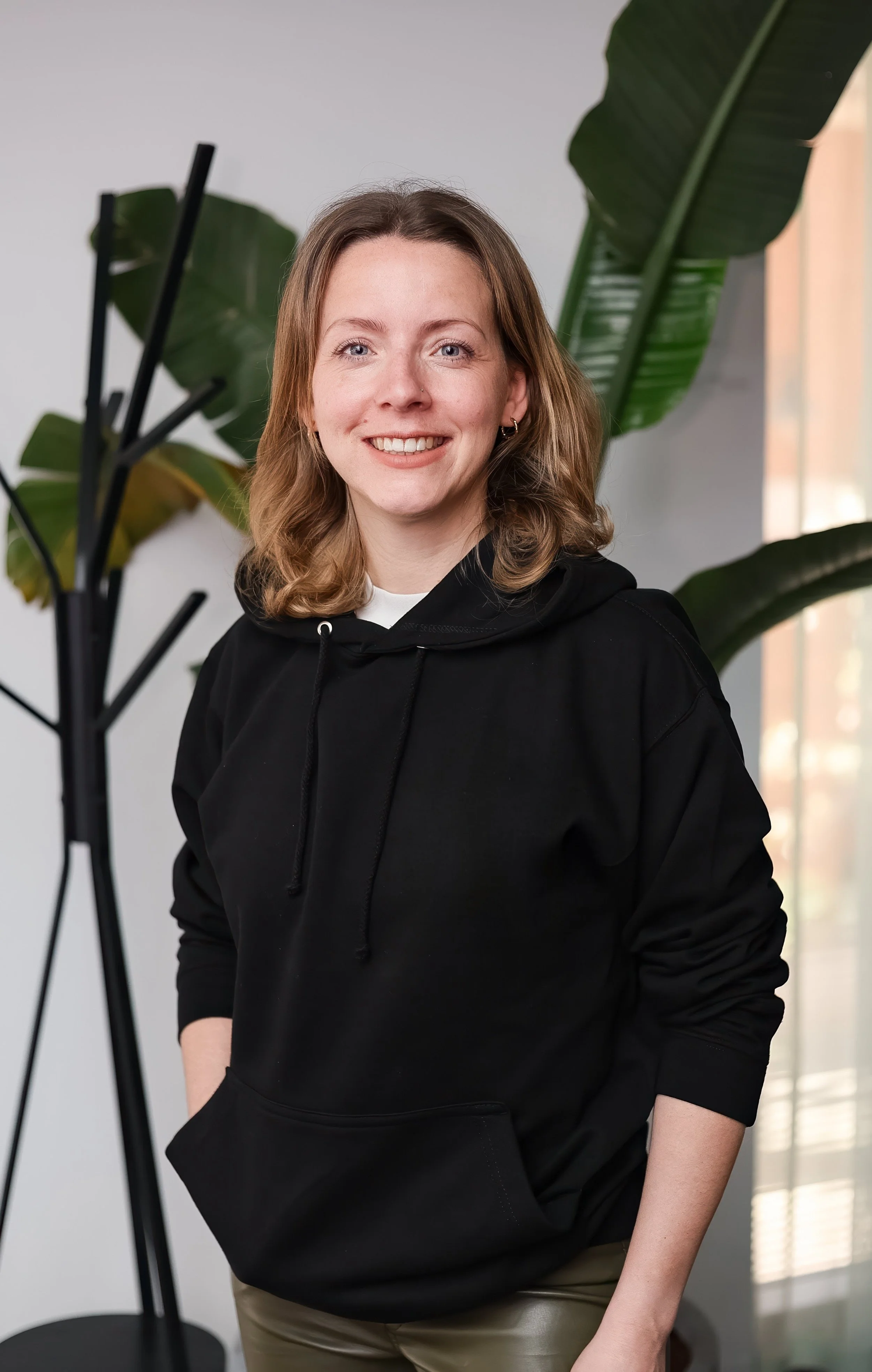 A smiling woman with shoulder-length blonde hair, wearing a black hoodie and beige pants, standing indoors near a large plant with broad green leaves, with a window in the background.