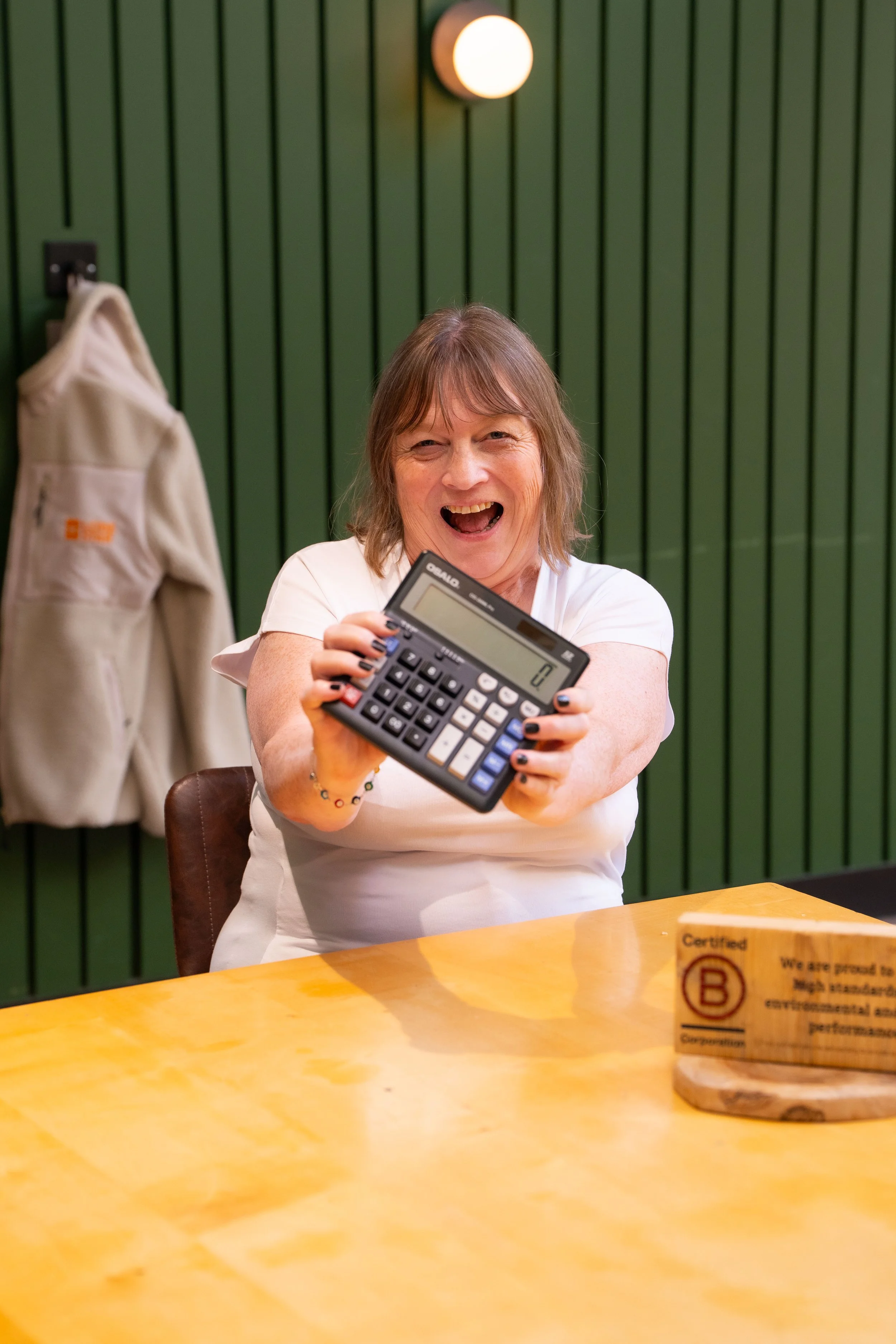 A smiling woman with shoulder-length brown hair, wearing a white shirt, holding a calculator, sitting at a yellow table in a room with green paneled walls and a round wall light.