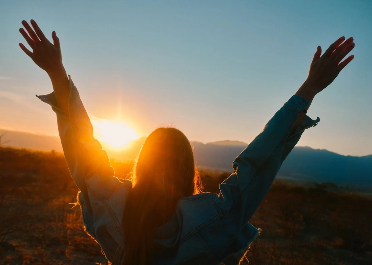 Person with long hair and denim jacket raising arms at sunset in a scenic outdoor landscape.