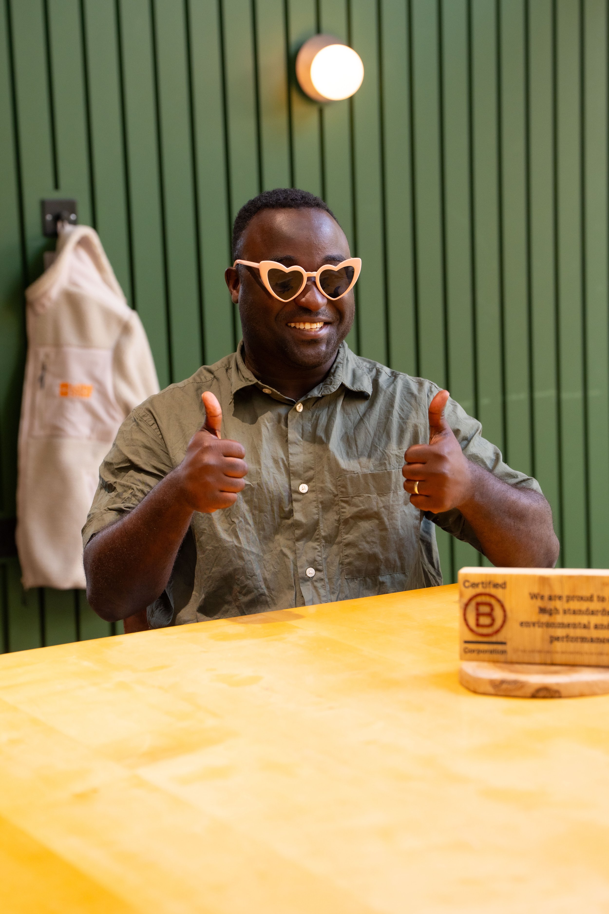 A man smiling and giving two thumbs up while wearing pink heart-shaped sunglasses, sitting at a table with a green wooden wall and a beige hanging bag in the background.