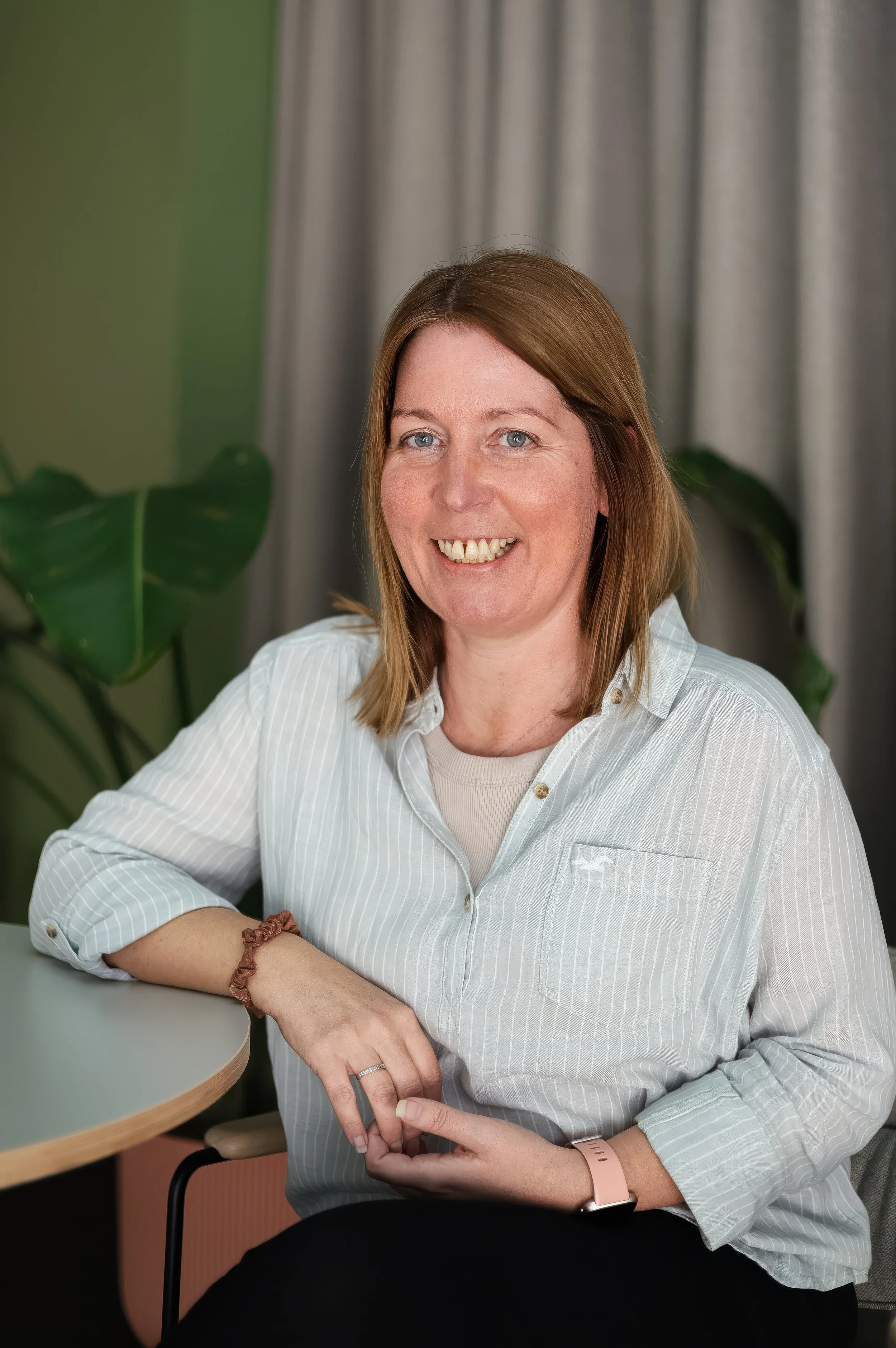 A woman with shoulder-length brown hair, blue eyes, and a fair complexion, smiling and sitting at a table in a room with gray curtains and green plants. She is wearing a light gray, striped, button-down shirt and has a pink watch and a brown hair tie bracelet on her wrist.