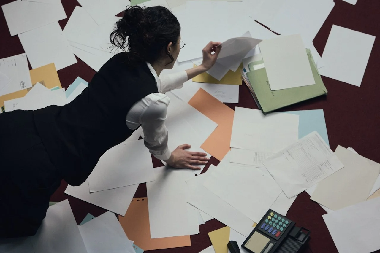 Woman lying on the floor amidst scattered papers, files, and a calculator.