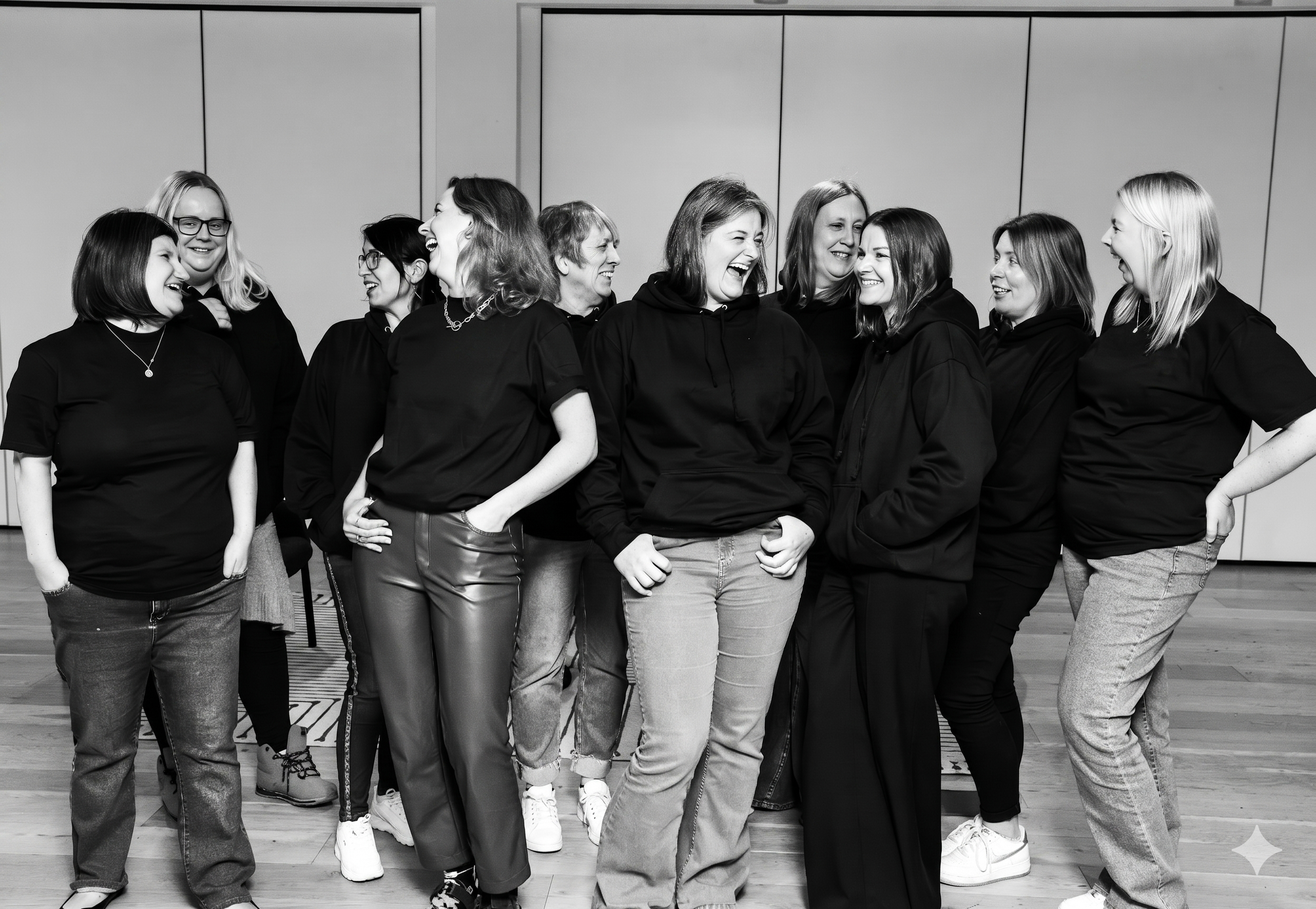 A group of women standing together, laughing and smiling in a social setting, with a plain wall background.