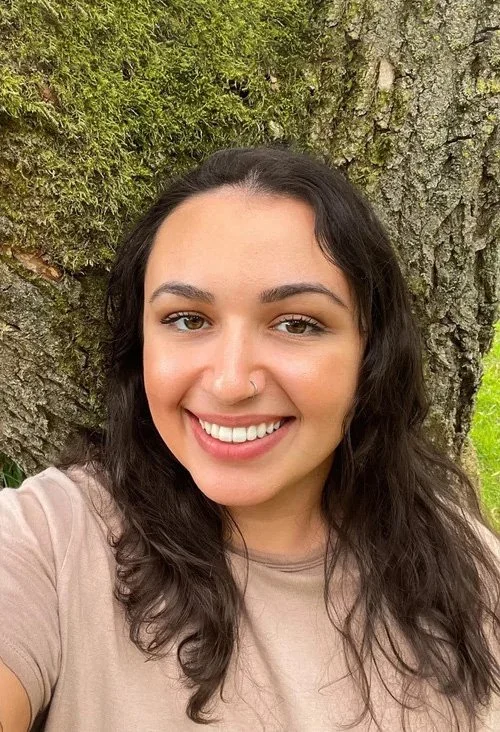 A young woman with long, wavy dark hair smiling in front of a large tree trunk with green moss and leaves.