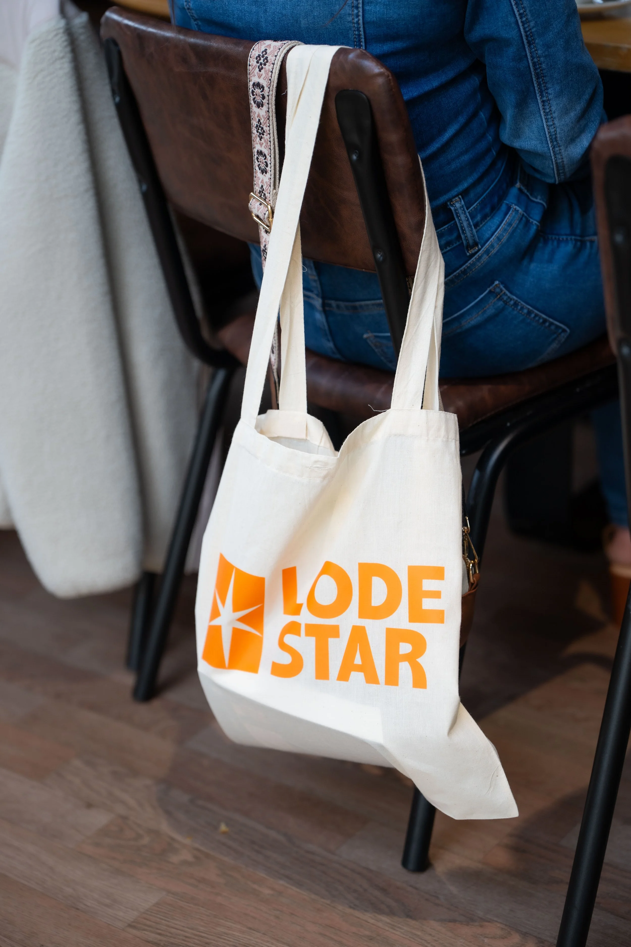 A beige tote bag with orange text that reads "LONE STAR" hanging on a chair in a room with wooden flooring and a person in blue jeans sitting nearby.