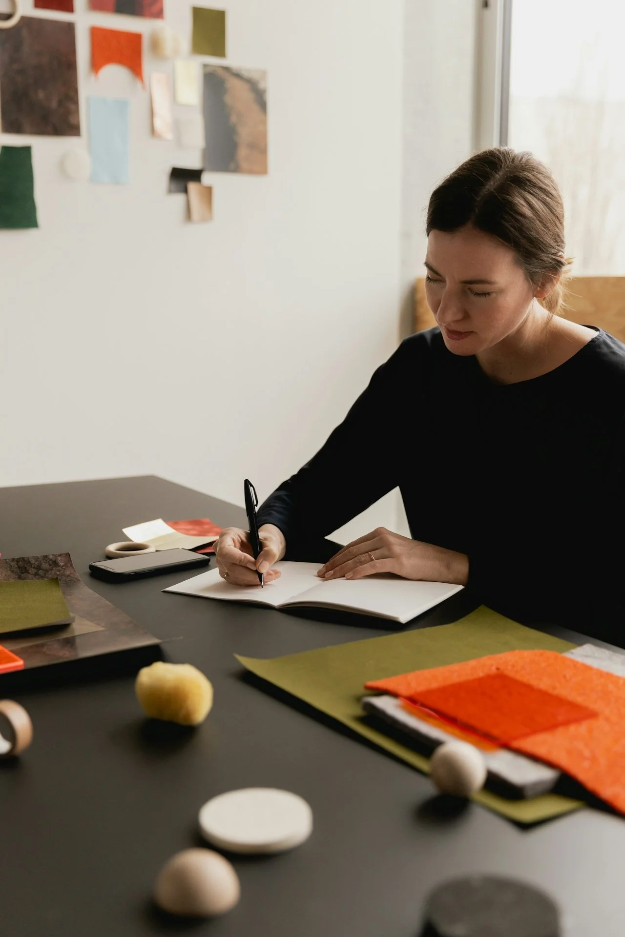 A woman sits at a desk, writing in a notebook, with various craft supplies and a smartphone on the table. The background features a white wall with colorful, abstract artwork.