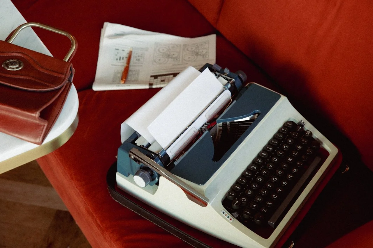 A vintage typewriter on a red table with papers, a brown leather purse, and a magazine or sheet of paper with diagrams and a pen.