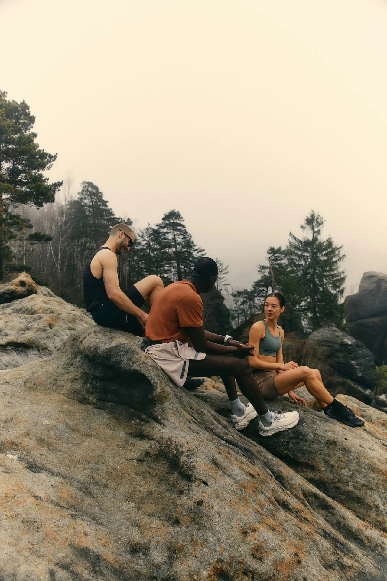 Three friends sitting on large rocks outdoors surrounded by trees, relaxing and conversing.