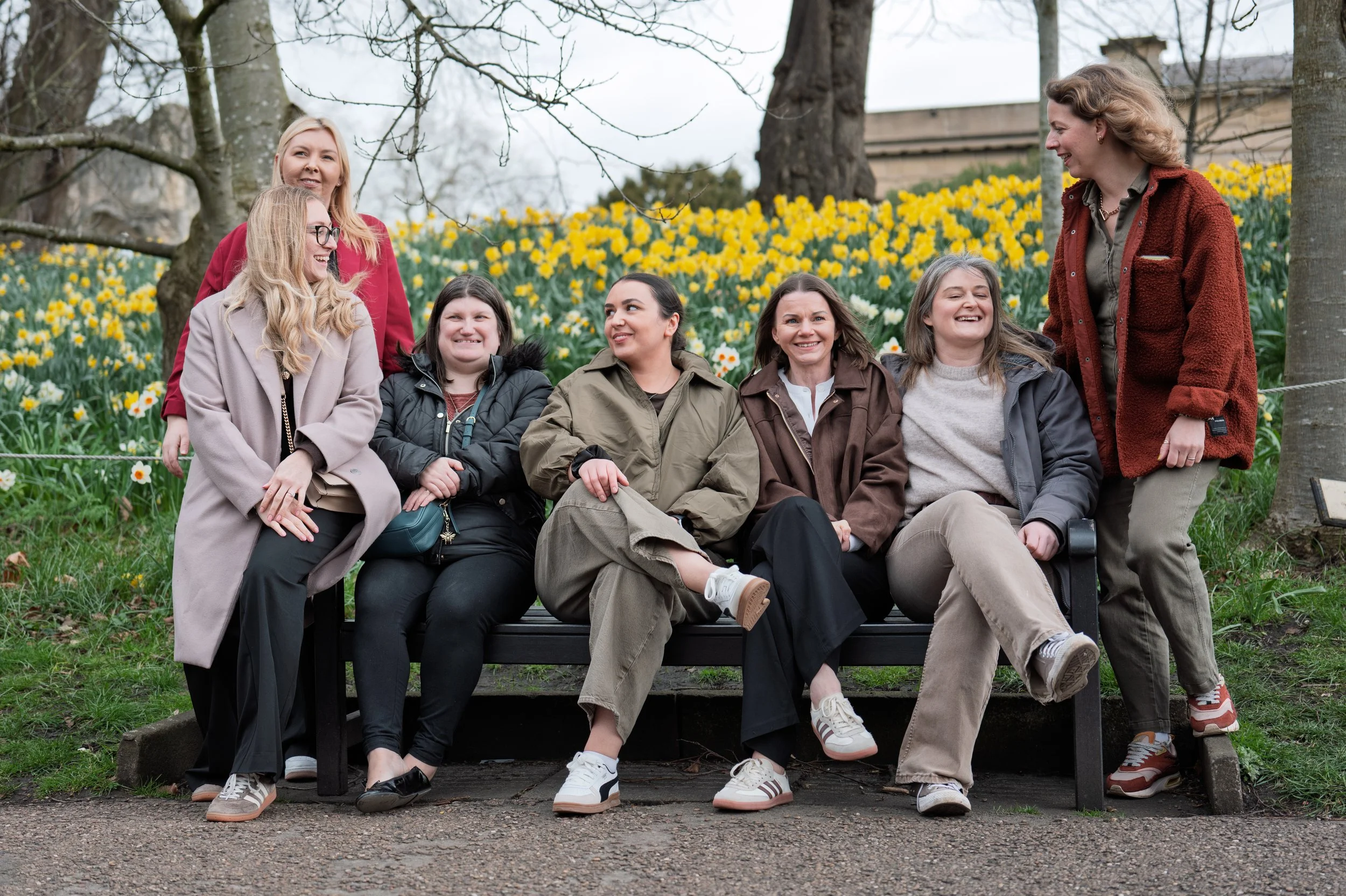 Group of seven women sitting and standing on a park bench surrounded by trees and yellow flowers, outdoors in spring.