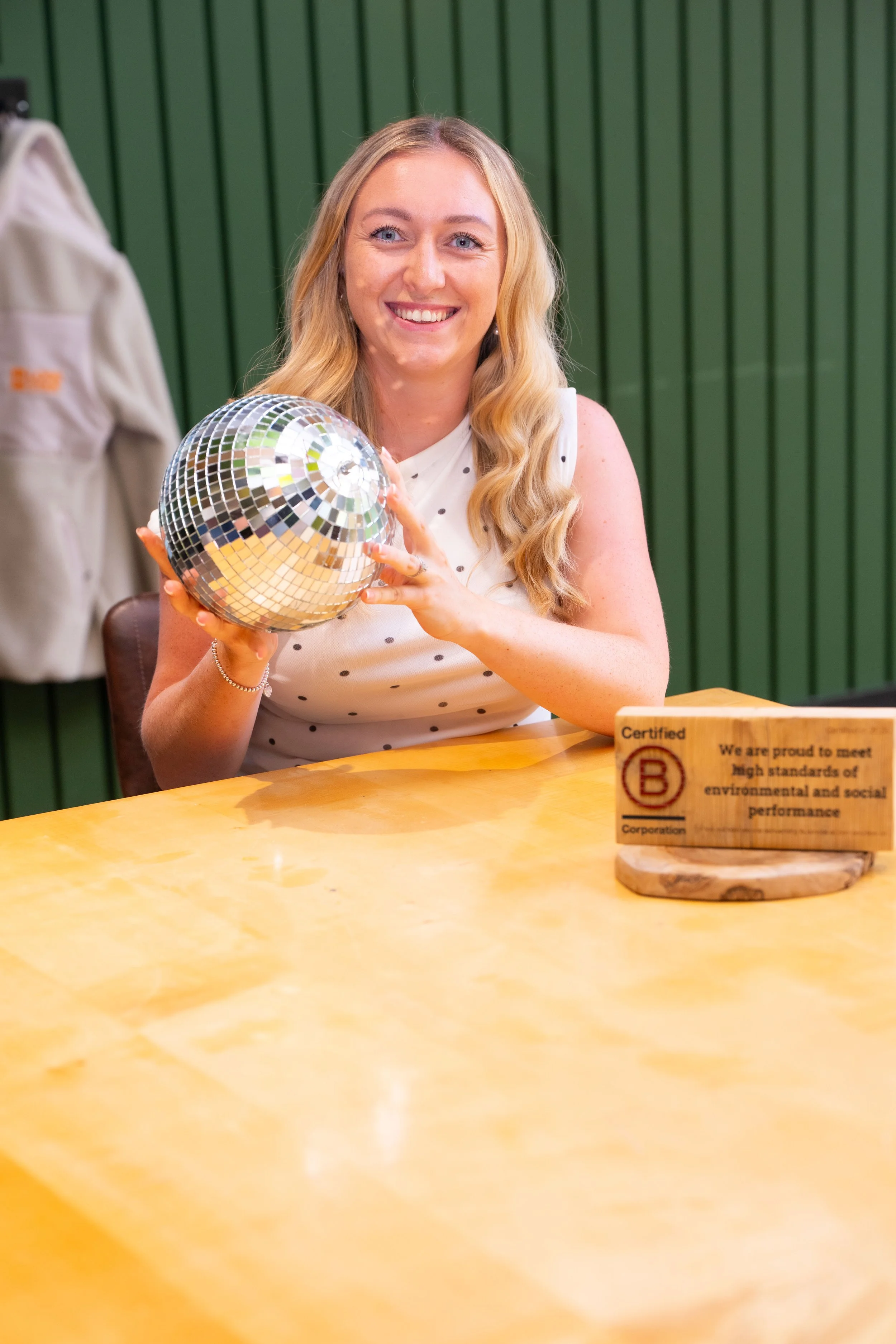 A woman with long wavy blonde hair smiling while holding a shiny disco ball in a room with green wood-paneled walls and a wooden table in front.