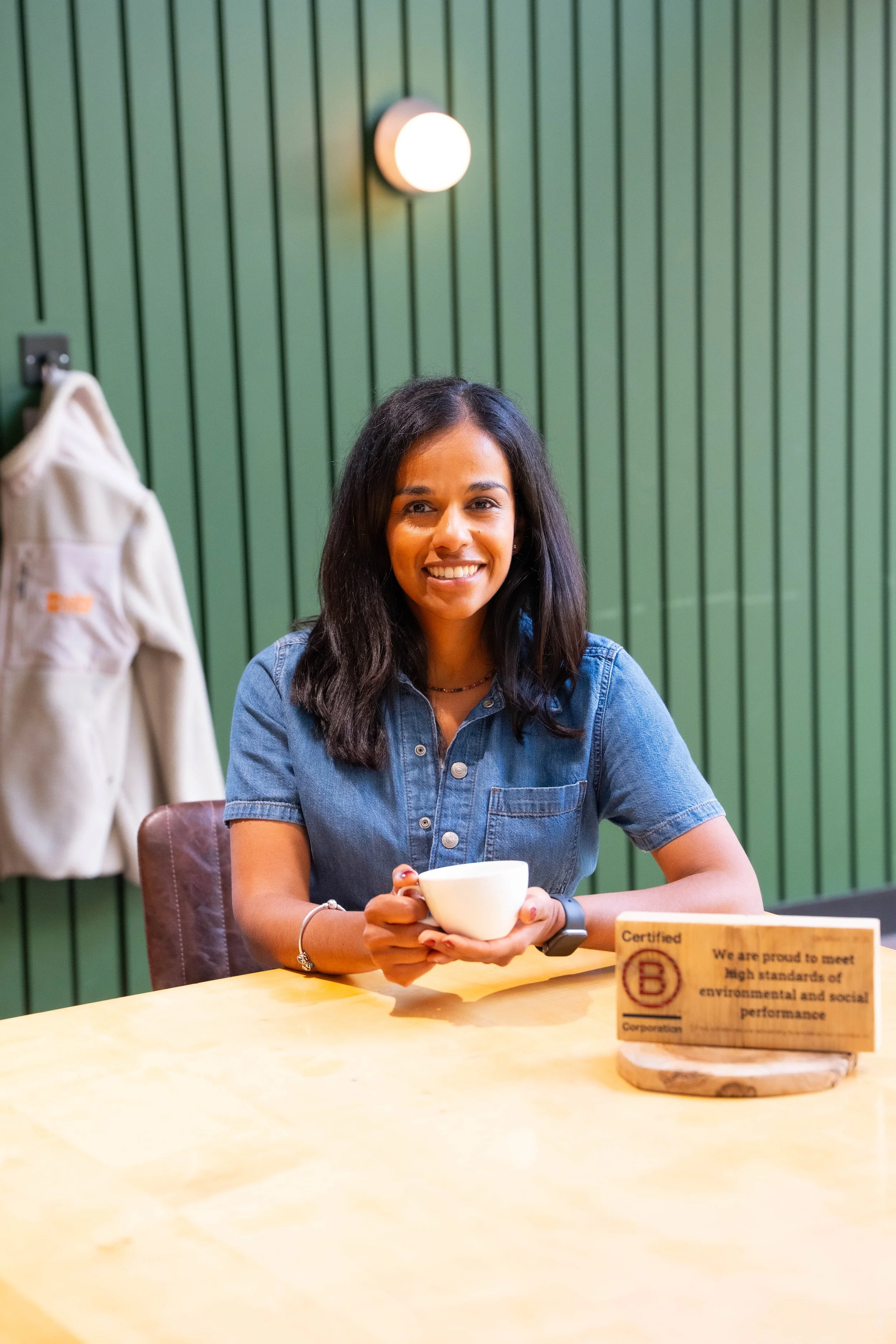 A smiling woman with long dark hair, wearing a denim shirt, holds a white mug and sits at a light wooden table. The background features a green paneled wall with a circular light fixture, and a beige bag hangs on a hook to the left. There is a small wooden sign on the table with text about environmental and social performance.