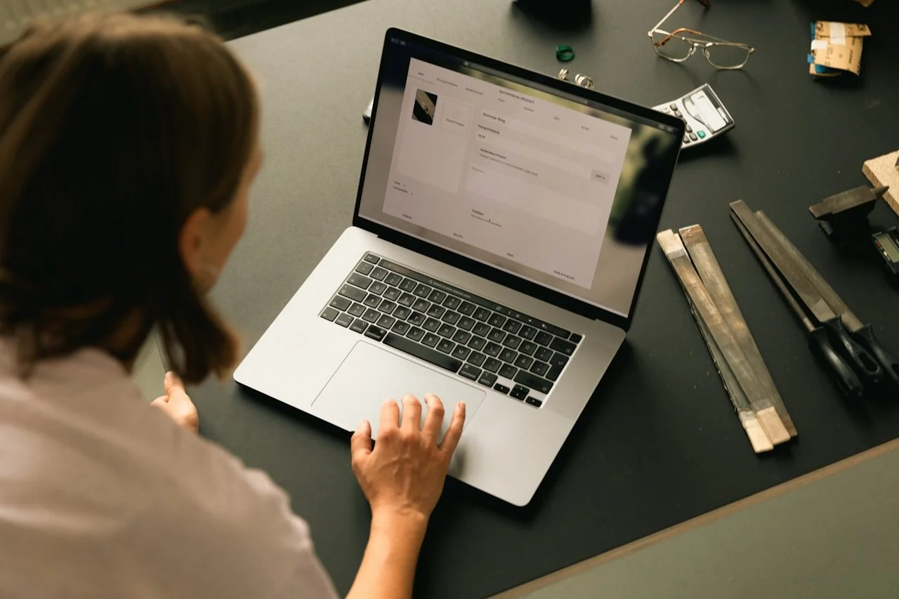 A woman sitting at a workbench using a laptop, with tools like rulers, scissors, and a calculator on the desk.