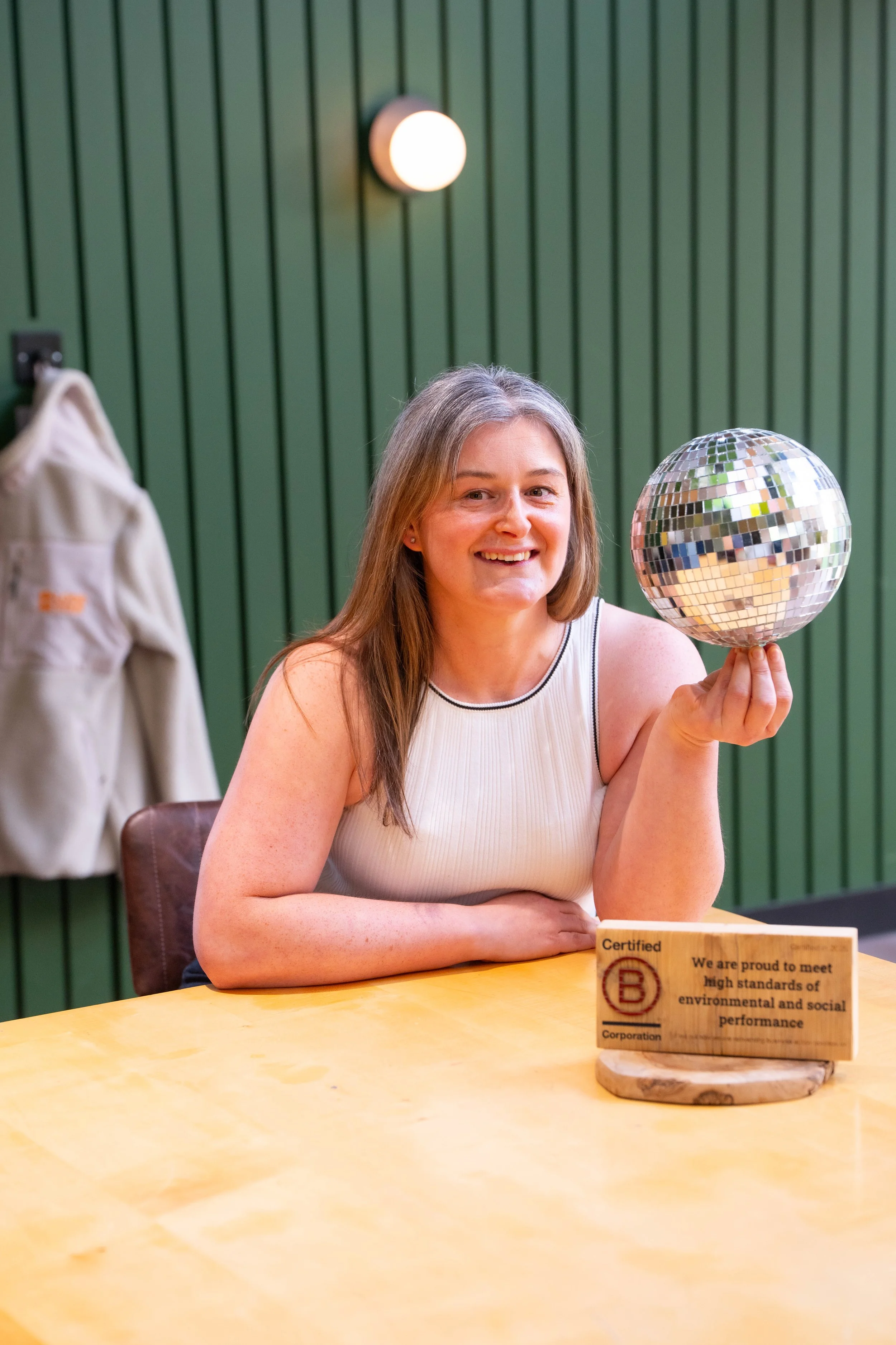 A woman with long gray hair smiling and holding a small disco ball with her right hand, sitting at a wooden table with a certification plaque in front of her. The background has green vertical wood paneling and a round light fixture.