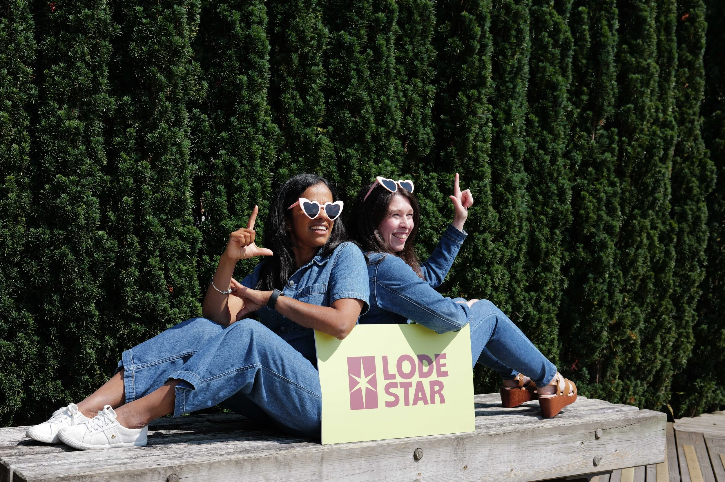 Two women wearing heart-shaped sunglasses, sitting on a wooden platform, holding a sign that says "LOVE STAR," in front of a hedge, smiling and pointing upwards.