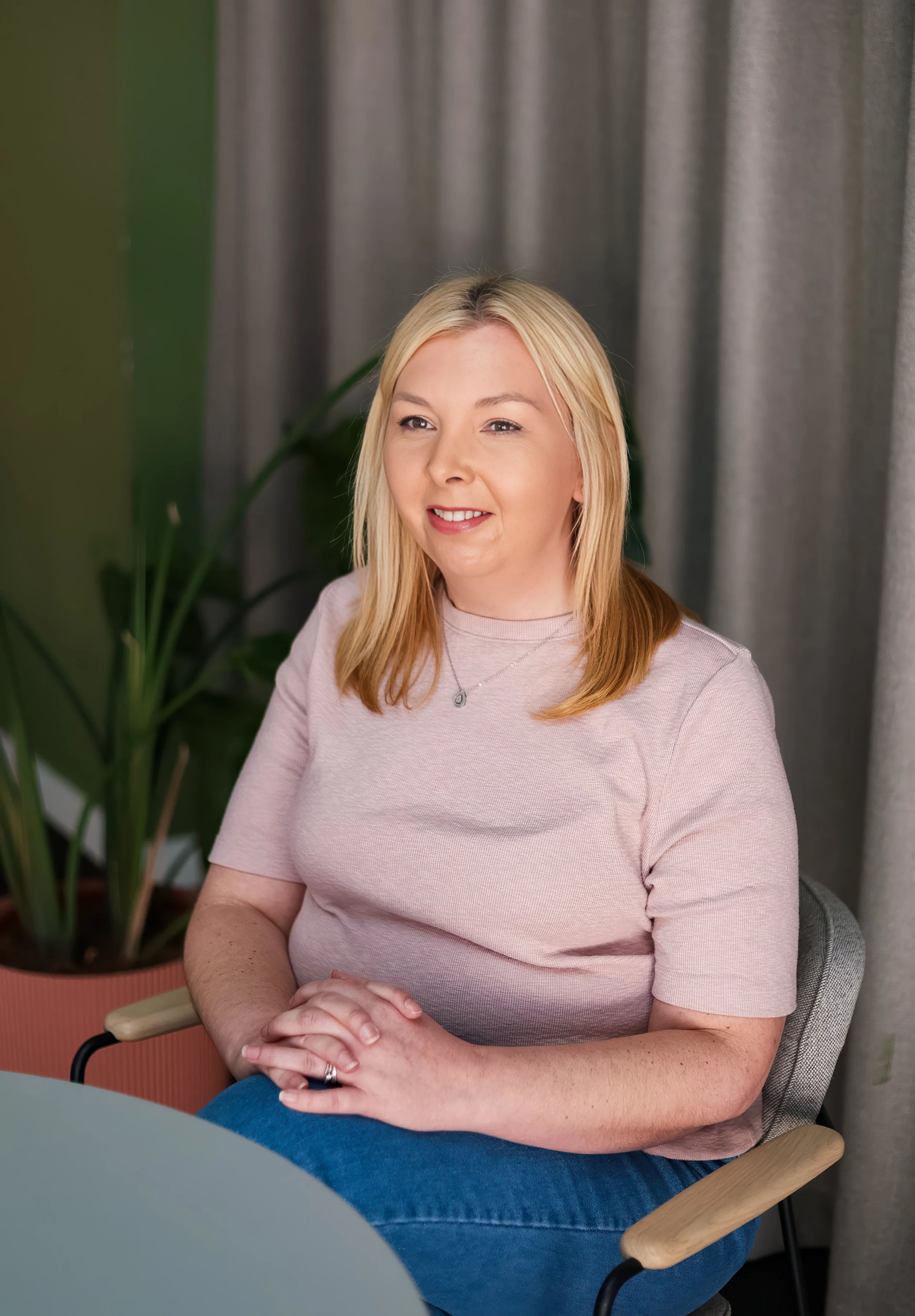 A blonde woman sits at a table with her hands folded, wearing a light pink t-shirt and a necklace, in front of a gray curtain and a green plant.