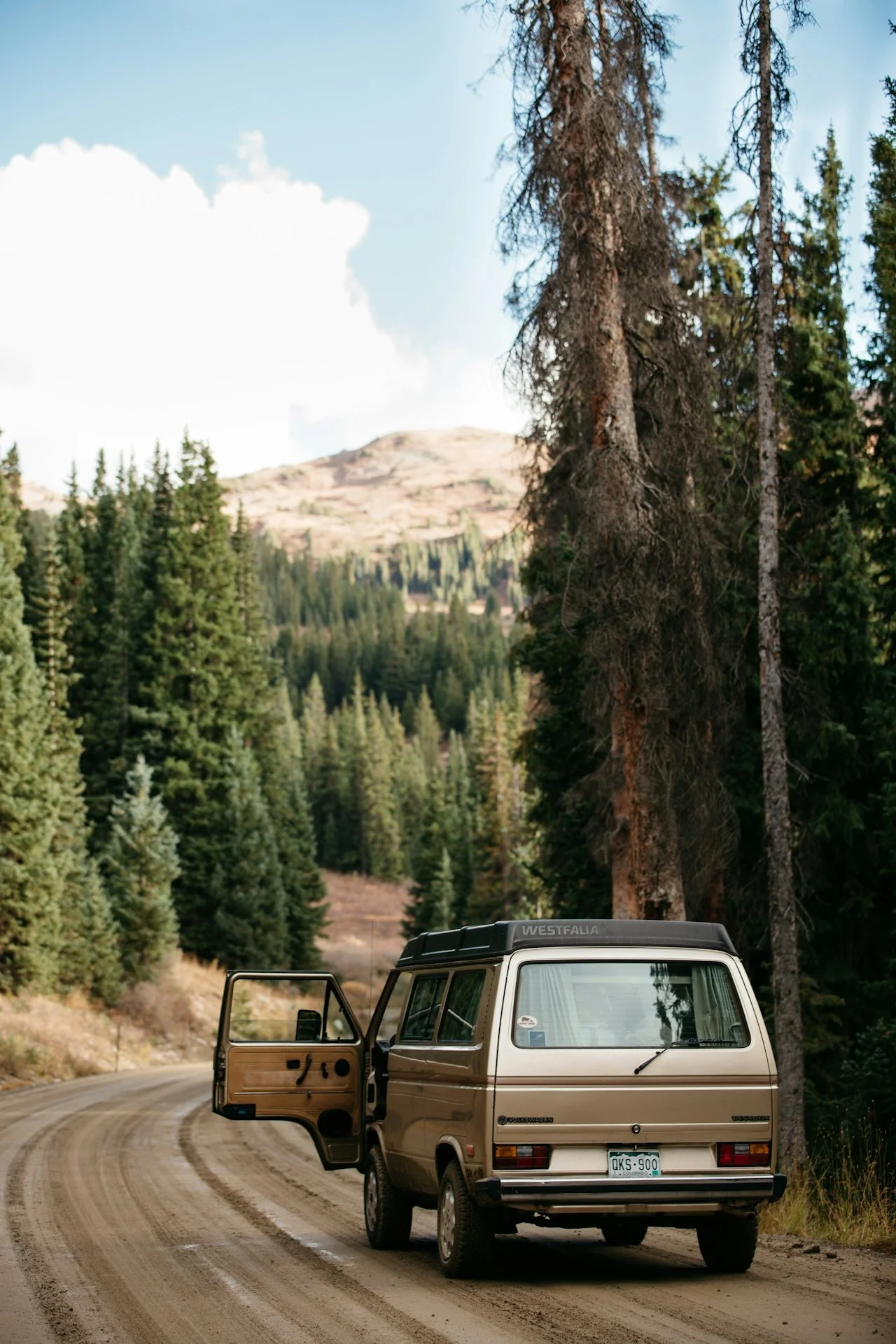 A vintage beige van parked on a winding dirt road in a forested mountain area, with the driver's side door open.