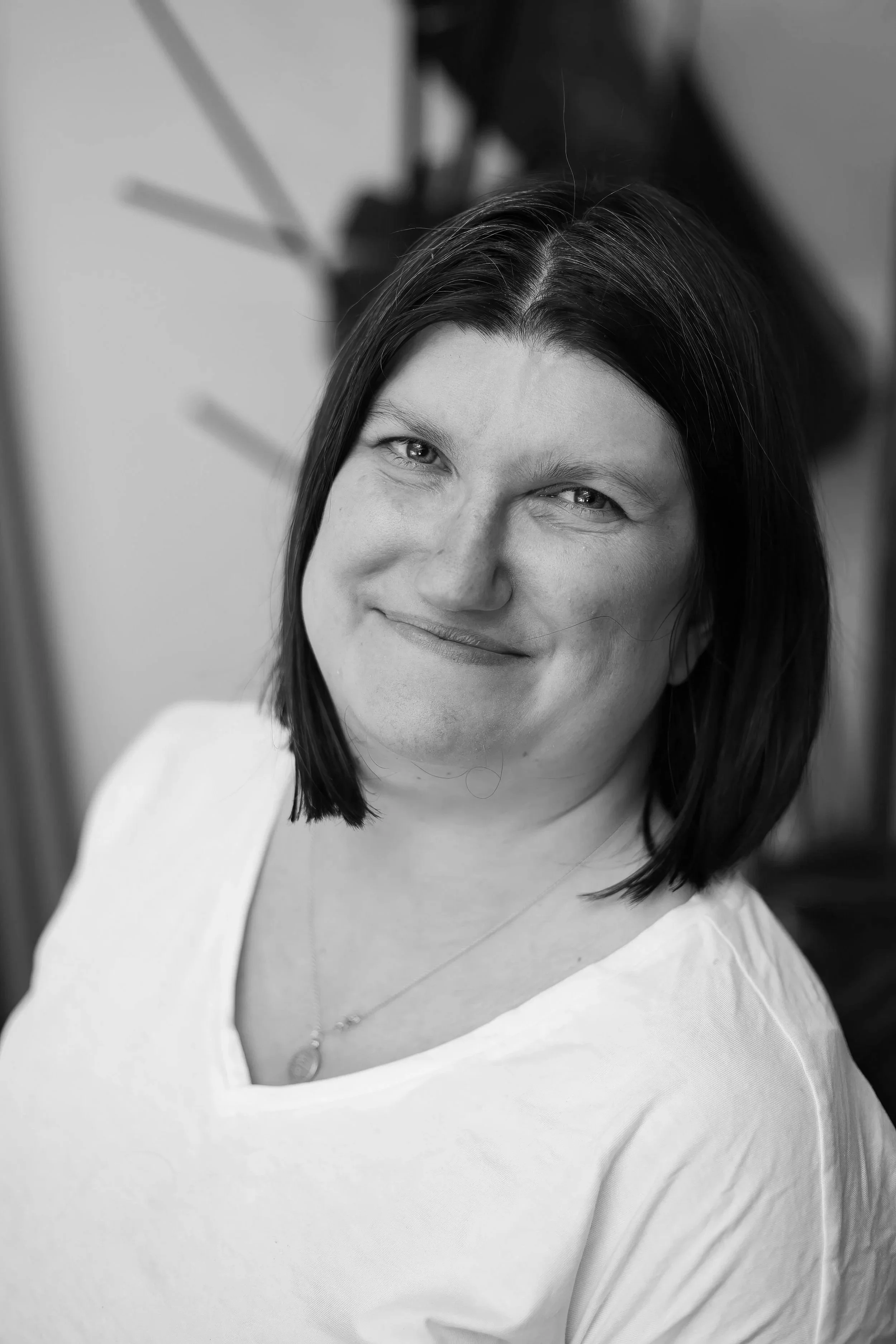 Black and white close-up of a smiling woman with dark hair, wearing a white top and necklace, with a blurred background.