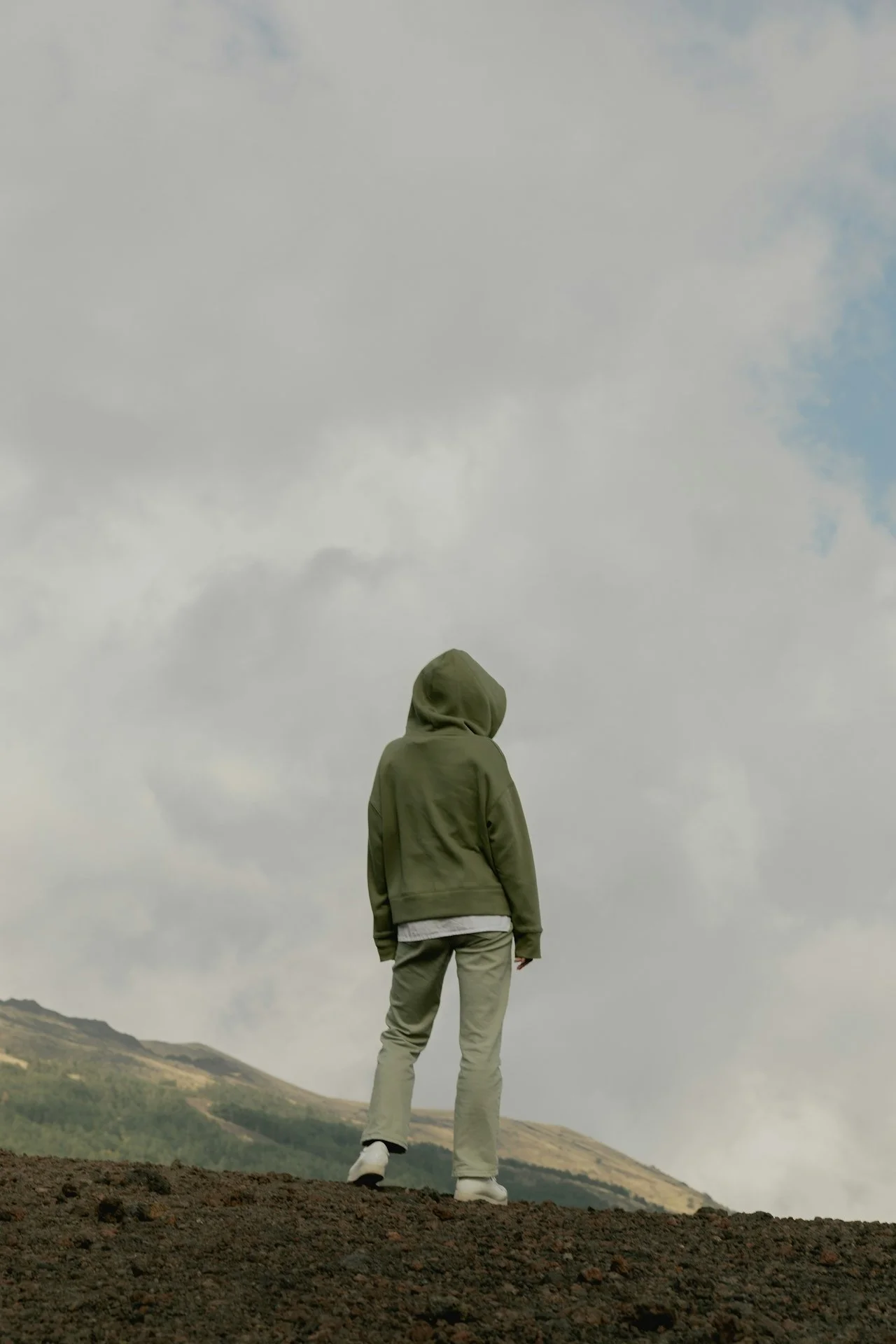 Person wearing a green hoodie and beige pants standing outdoors on a cloudy day, looking at the cloudy sky with hills in the background.