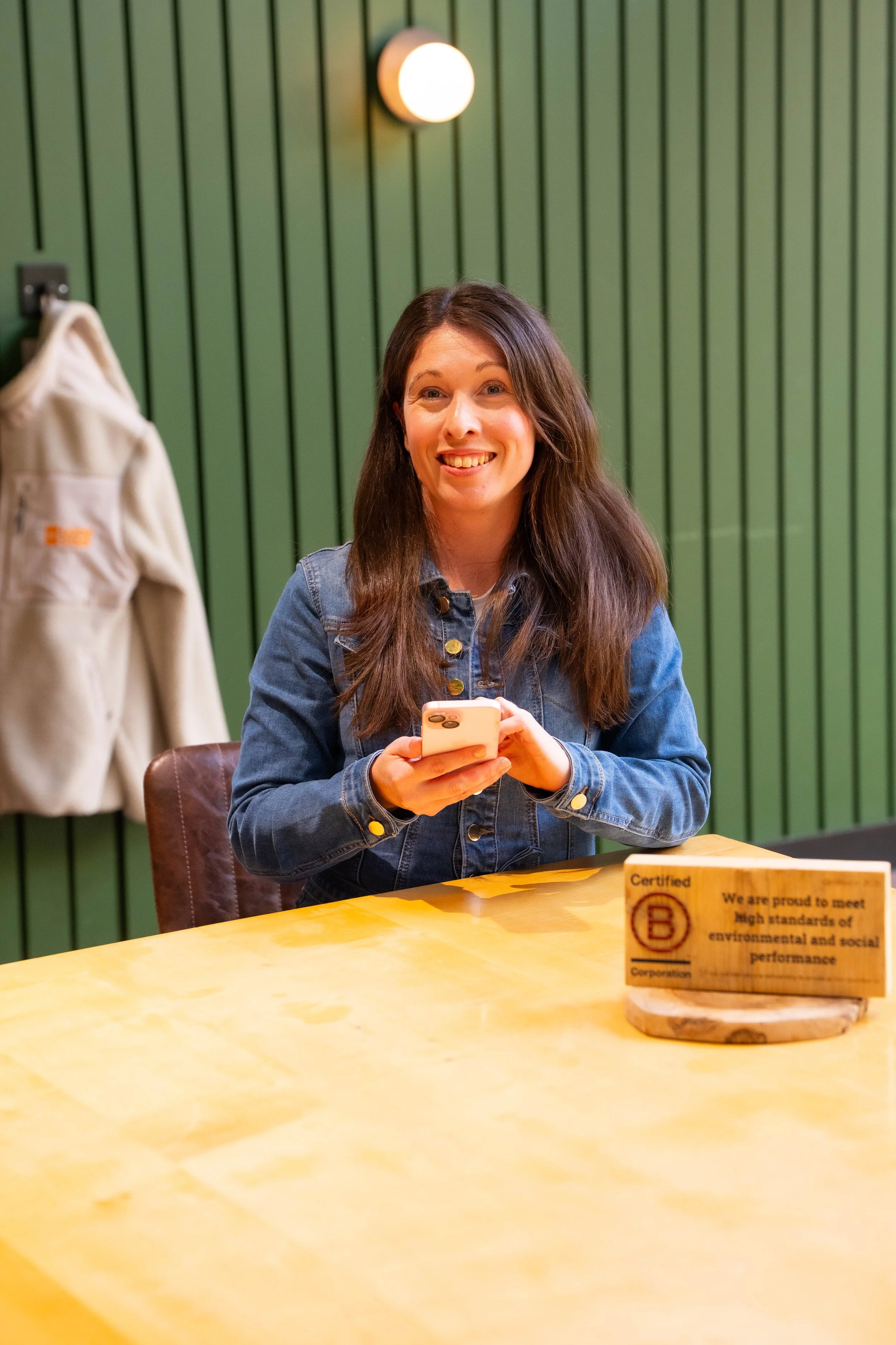 A woman with long brown hair, wearing a denim jacket, sitting at a yellow table in a green-paneled room, smiling while holding a smartphone. A beige coat hangs to her left, and a wooden sign with text is on the table.