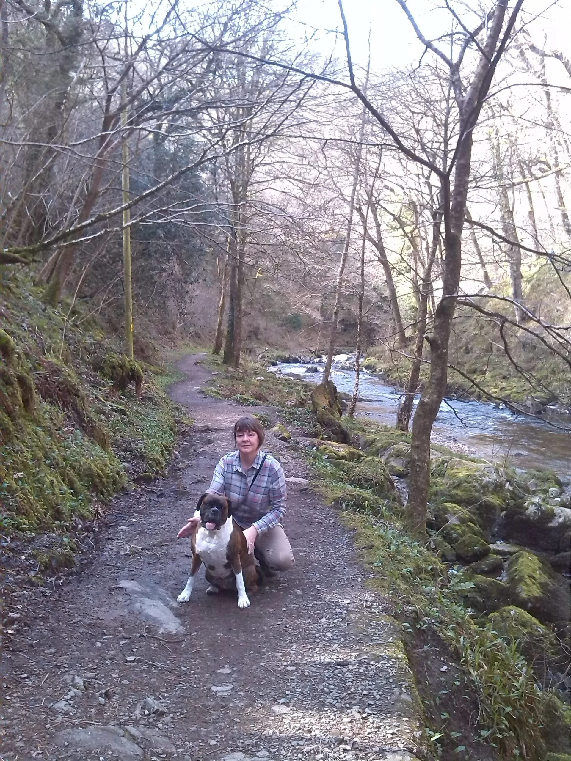 A woman in a plaid shirt kneeling on a dirt trail, holding a dog, beside a creek with trees in a forested area.