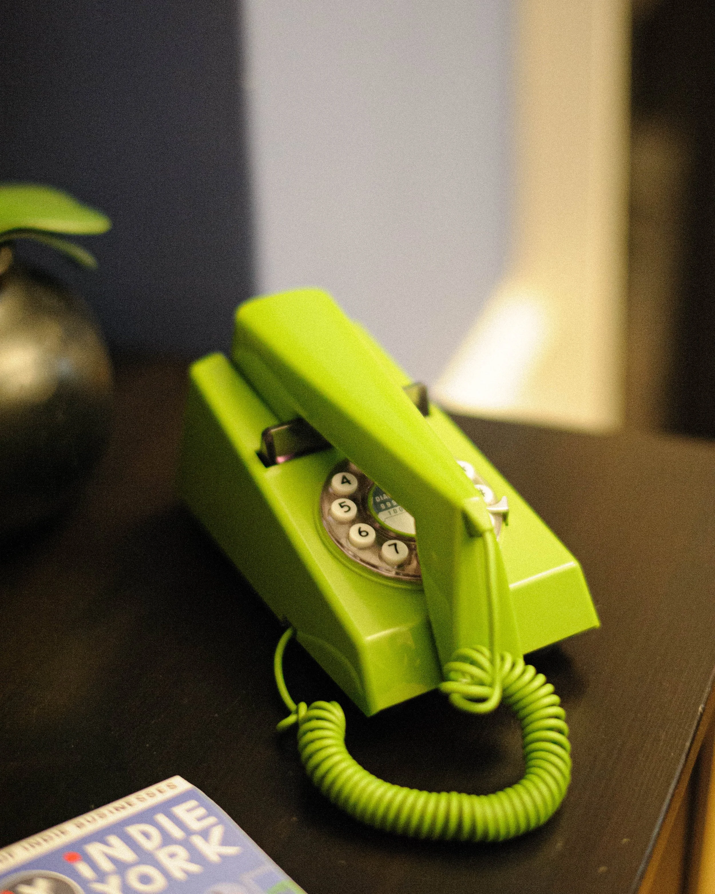 A bright green rotary dial telephone with its cord coiled below, sitting on a dark surface. Part of a magazine or booklet with the words "THE PEOPLE'S WORK" visible in the foreground.