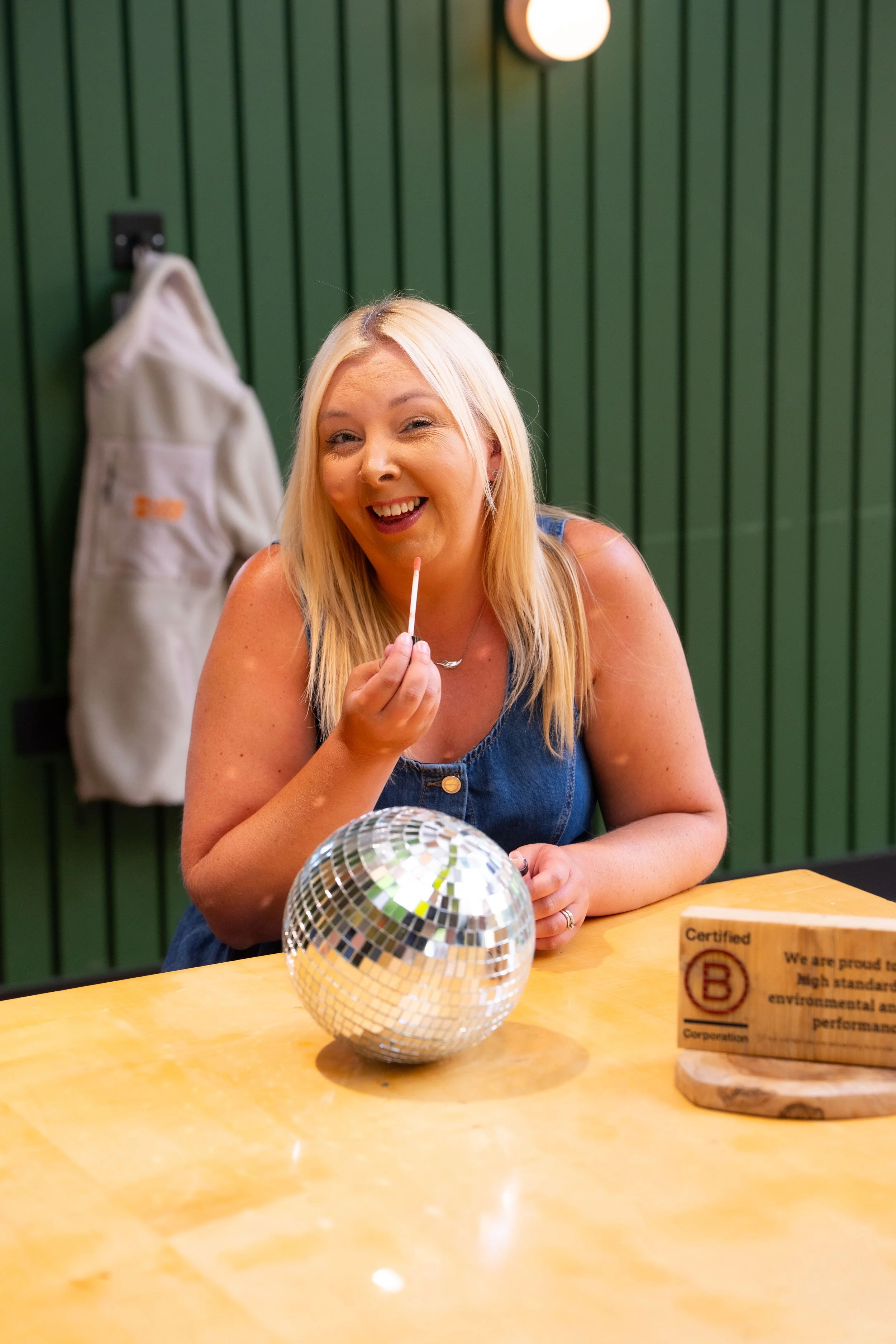 A woman with blonde hair smiling and winking while holding a white stick, sitting at a yellow table with a disco ball in front of her, in front of green wall panels and a wall light.