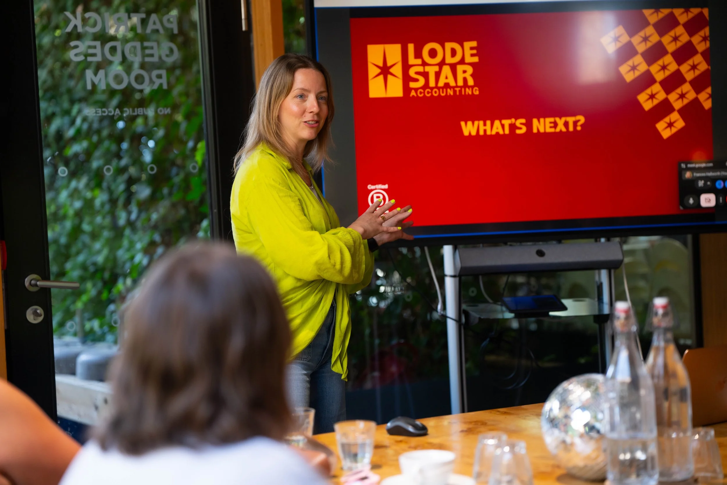 A woman with shoulder-length blond hair wearing a yellow jacket is presenting in front of a red screen that reads 'Lode Star Accounting' and 'What's Next?'. She is in a room with large windows showing green foliage outside. There are audience members and a table with drinks and snacks in the foreground.